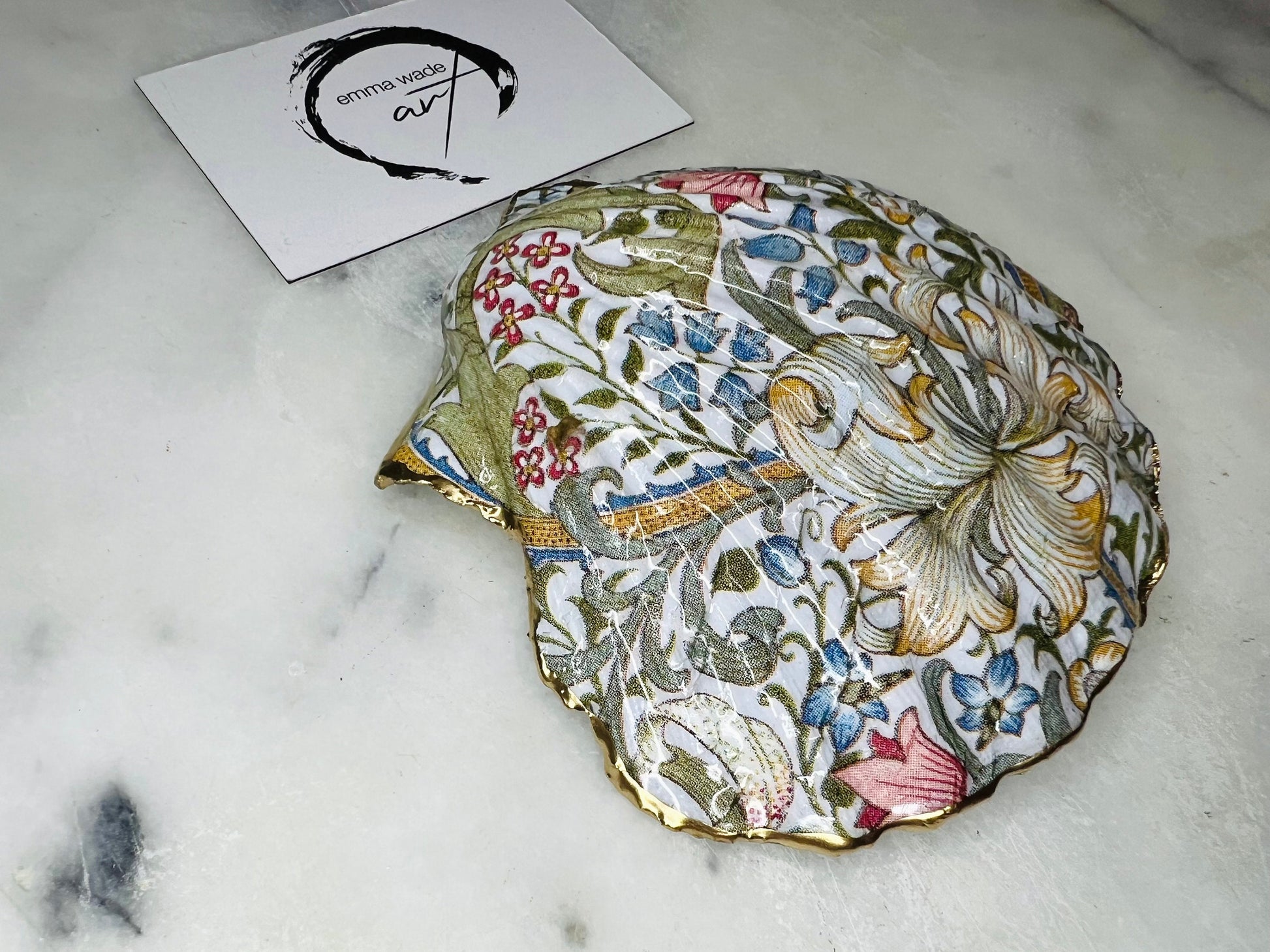 Handcrafted Dorset scallop shell trinket dish featuring William Morris Golden Lily floral design. Natural shell edged with hand applied gold leaf and sealed with glossy resin. Heritage floral jewellery holder displayed with rings on marble surface. Elegant coastal home decor gift.