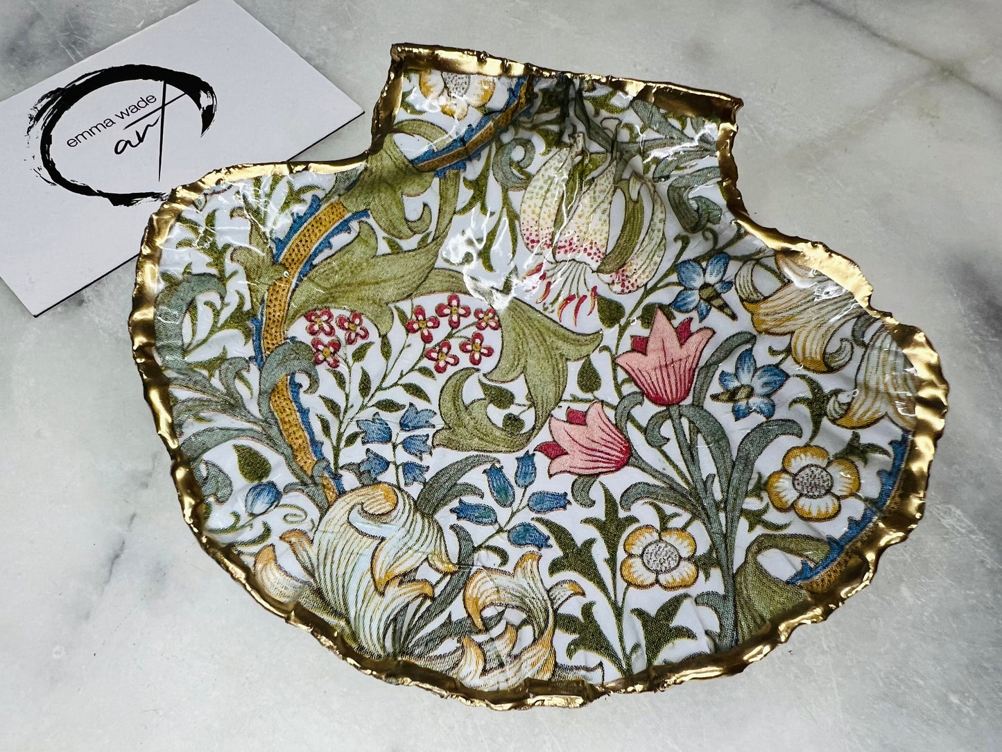 Handcrafted Dorset scallop shell trinket dish featuring William Morris Golden Lily floral design. Natural shell edged with hand applied gold leaf and sealed with glossy resin. Heritage floral jewellery holder displayed with rings on marble surface. Elegant coastal home decor gift.