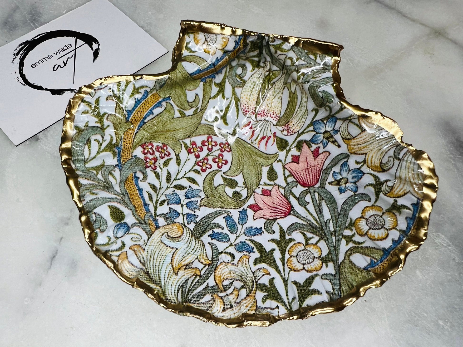 Handcrafted Dorset scallop shell trinket dish featuring William Morris Golden Lily floral design. Natural shell edged with hand applied gold leaf and sealed with glossy resin. Heritage floral jewellery holder displayed with rings on marble surface. Elegant coastal home decor gift.