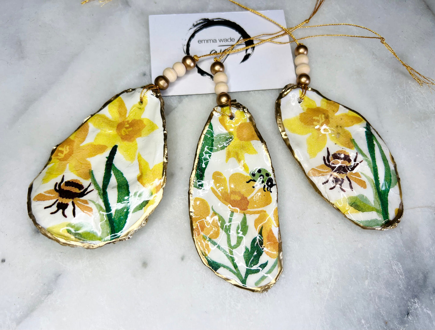 Handcrafted Dorset oyster shell hanging ornament featuring genuine Emma Bridgewater yellow spring floral napkin design with bright yellow flowers, green foliage, bumblebees and ladybirds. Finished with liquid gold leaf edging and sealed in glossy resin. Natural pearlescent reverse with gold detailing and gold hanging cord with decorative beads. Coastal seasonal home decor.