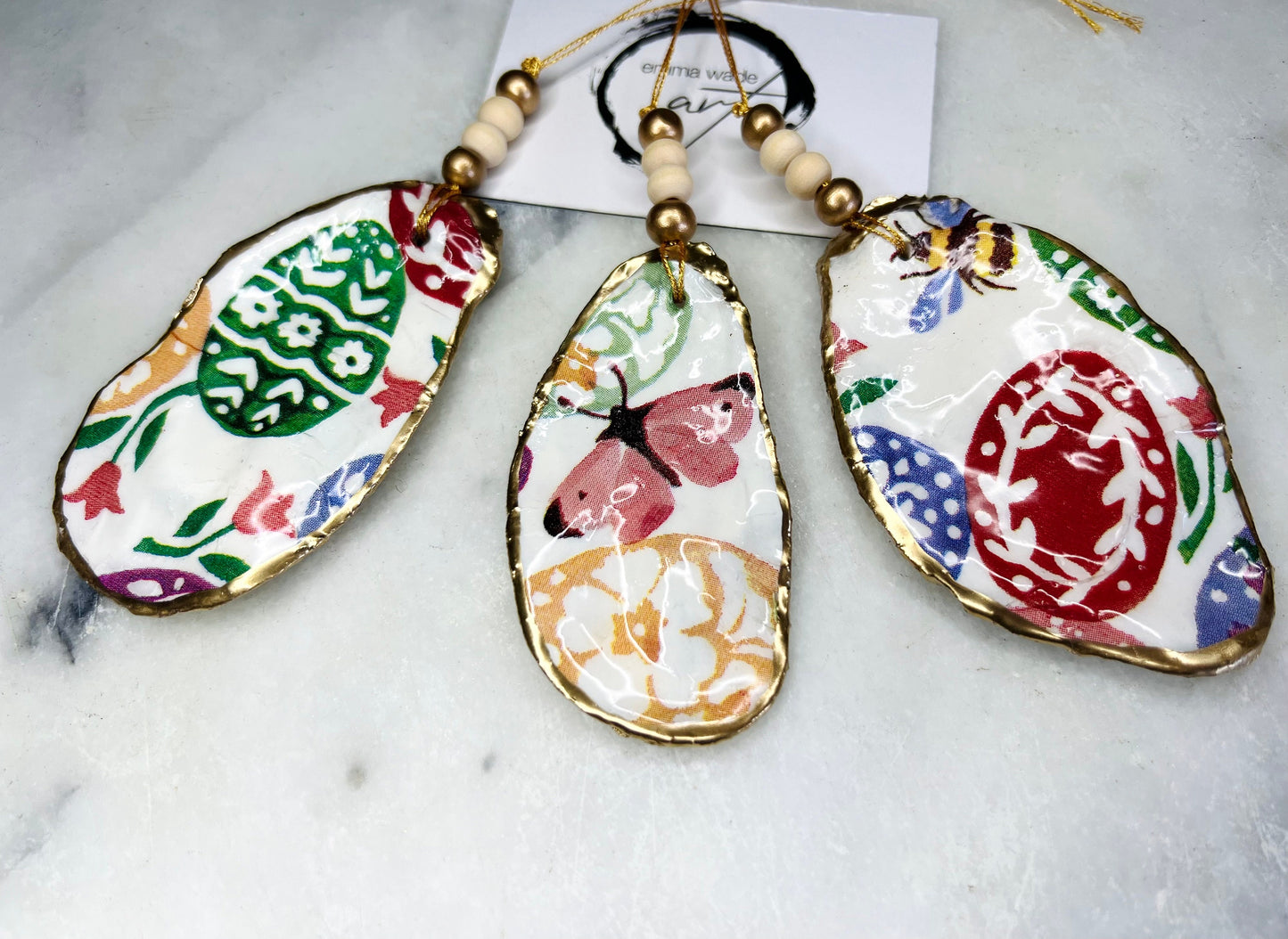 Handmade Dorset oyster shell ornaments featuring Emma Bridgewater bee, butterfly, and egg designs. Each shell has a natural pearl finish, gilded with liquid gold leaf on the rim and back, and sealed with glossy resin for shine and durability. Available individually or as a set of 3, with pre-drilled holes, gold hanging string, and decorative beads. Unique festive decorations for Christmas trees, seasonal home décor, or handmade coastal-inspired gifts for weddings, birthdays, or anniversaries.