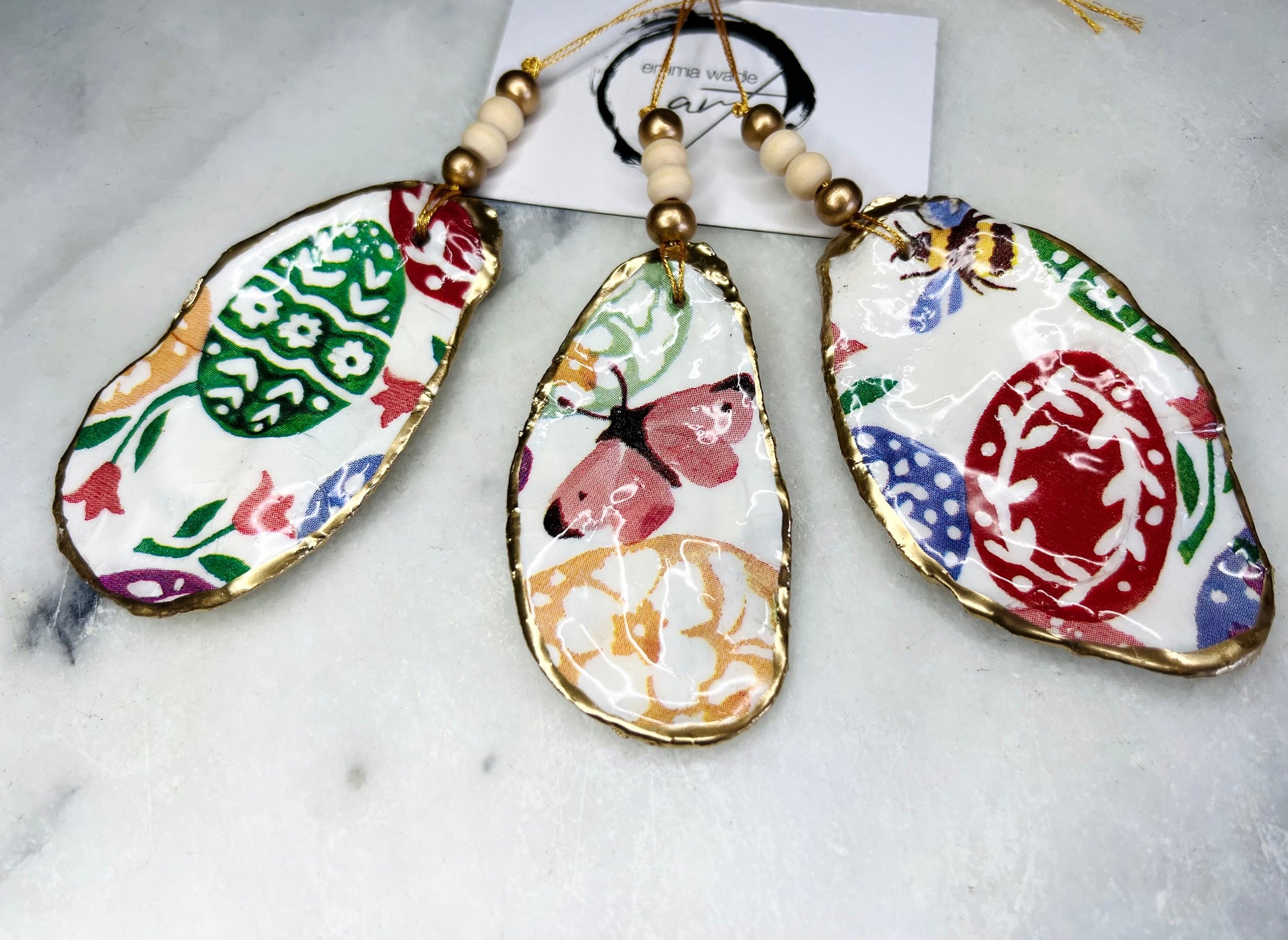 Handmade Dorset oyster shell ornaments featuring Emma Bridgewater bee, butterfly, and egg designs. Each shell has a natural pearl finish, gilded with liquid gold leaf on the rim and back, and sealed with glossy resin for shine and durability. Available individually or as a set of 3, with pre-drilled holes, gold hanging string, and decorative beads. Unique festive decorations for Christmas trees, seasonal home décor, or handmade coastal-inspired gifts for weddings, birthdays, or anniversaries.