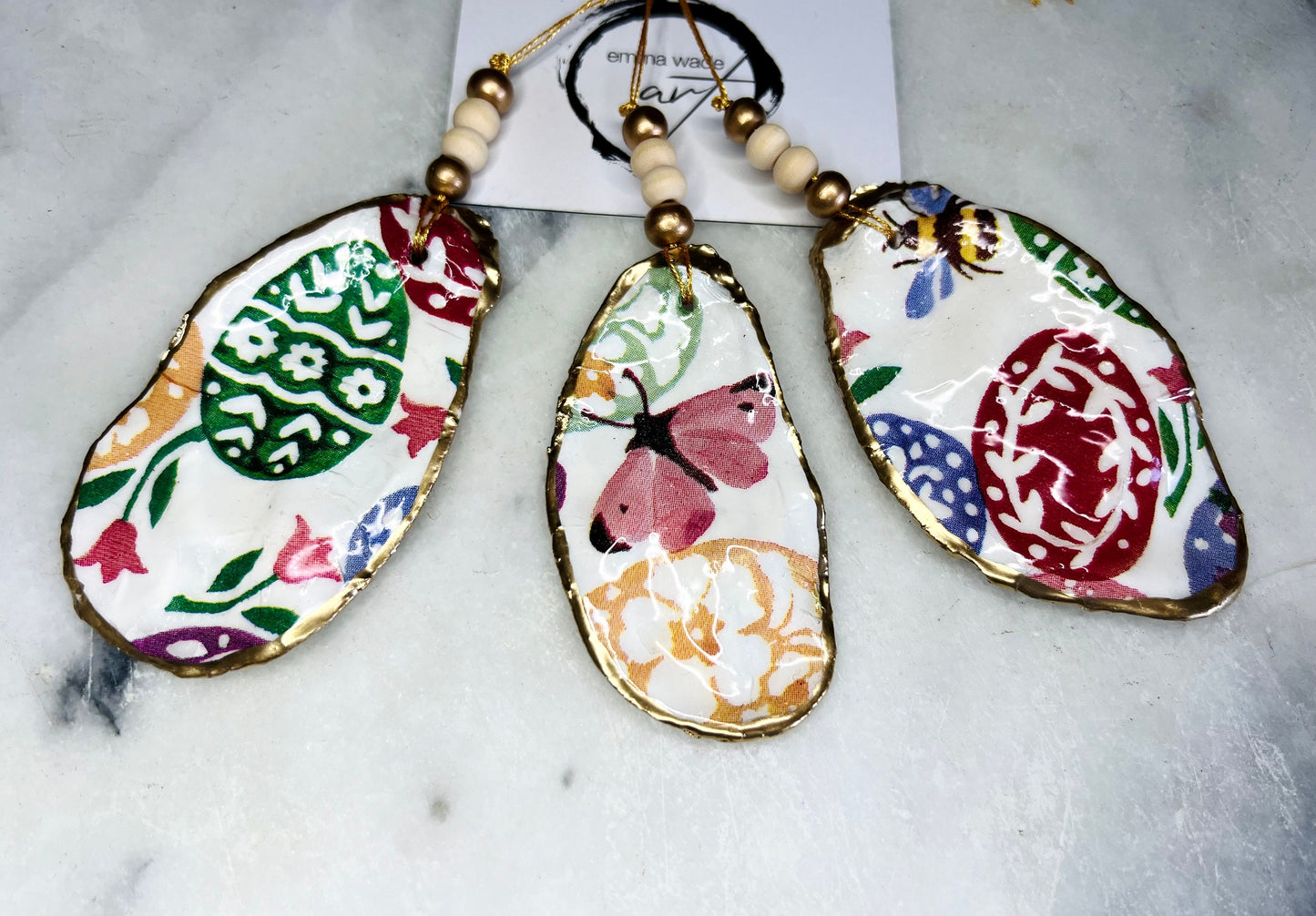 Handmade Dorset oyster shell ornaments featuring Emma Bridgewater bee, butterfly, and egg designs. Each shell has a natural pearl finish, gilded with liquid gold leaf on the rim and back, and sealed with glossy resin for shine and durability. Available individually or as a set of 3, with pre-drilled holes, gold hanging string, and decorative beads. Unique festive decorations for Christmas trees, seasonal home décor, or handmade coastal-inspired gifts for weddings, birthdays, or anniversaries.