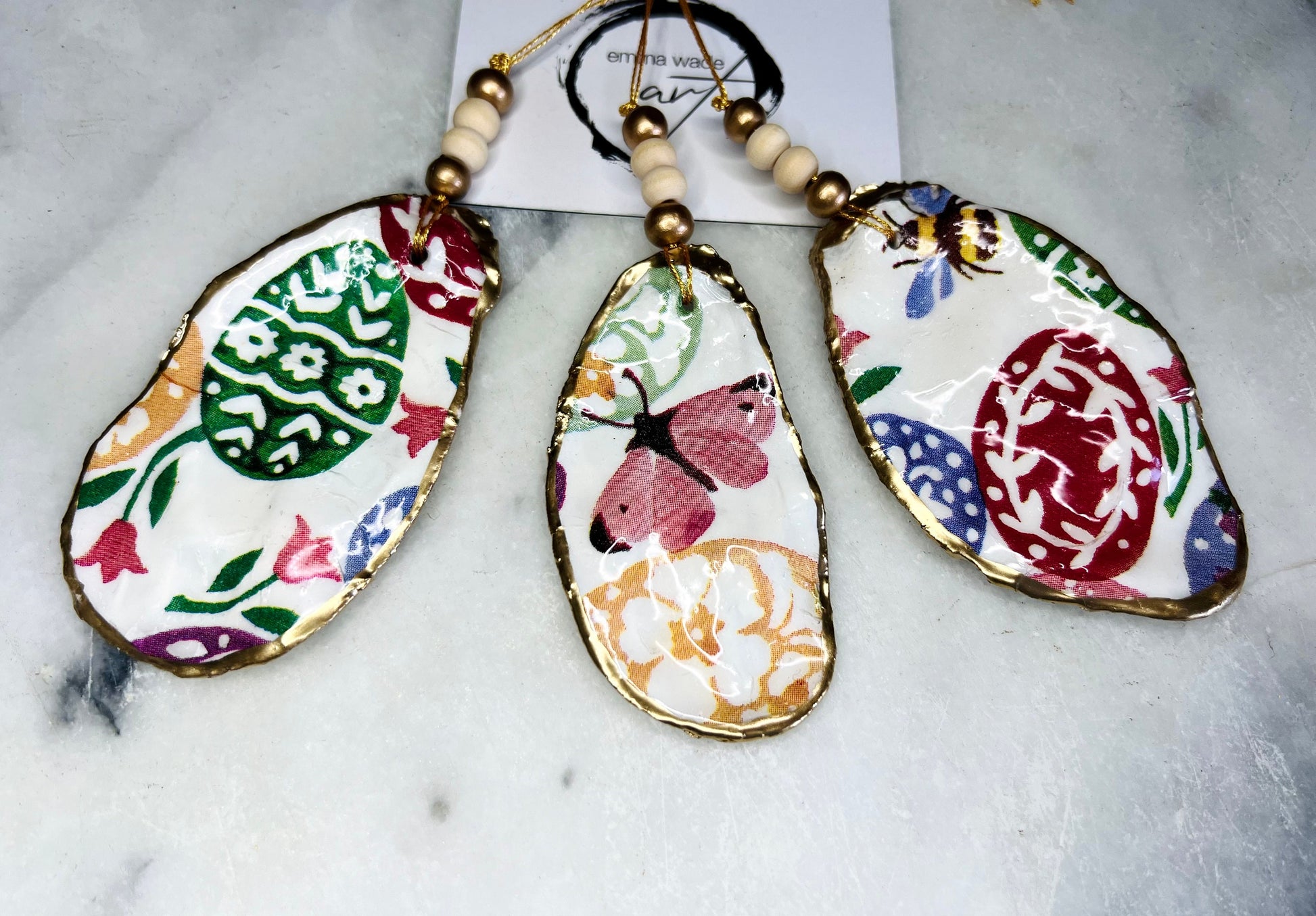 Handmade Dorset oyster shell ornaments featuring Emma Bridgewater bee, butterfly, and egg designs. Each shell has a natural pearl finish, gilded with liquid gold leaf on the rim and back, and sealed with glossy resin for shine and durability. Available individually or as a set of 3, with pre-drilled holes, gold hanging string, and decorative beads. Unique festive decorations for Christmas trees, seasonal home décor, or handmade coastal-inspired gifts for weddings, birthdays, or anniversaries.
