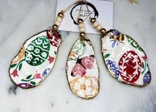 Handmade Dorset oyster shell ornaments featuring Emma Bridgewater bee, butterfly, and egg designs. Each shell has a natural pearl finish, gilded with liquid gold leaf on the rim and back, and sealed with glossy resin for shine and durability. Available individually or as a set of 3, with pre-drilled holes, gold hanging string, and decorative beads. Unique festive decorations for Christmas trees, seasonal home décor, or handmade coastal-inspired gifts for weddings, birthdays, or anniversaries.