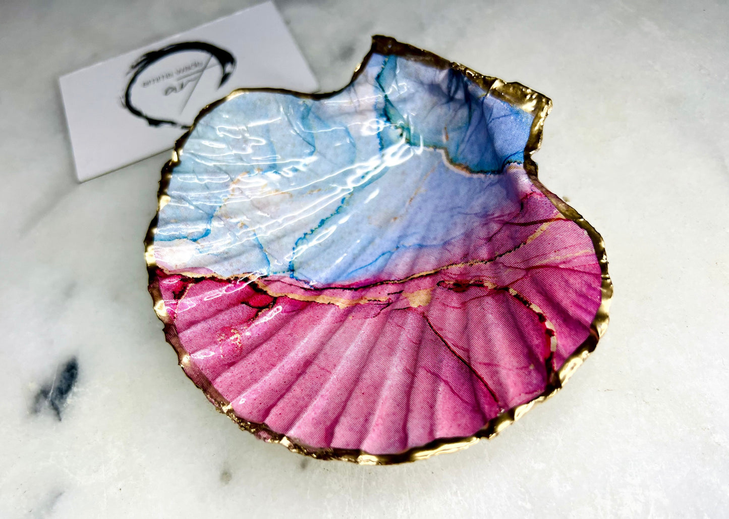 A handmade scallop shell trinket dish painted in soft pink and pale blue tones in a flowing, watercolor-style pattern, finished with a gold leaf edge and sealed with a glossy clear resin. The natural shell shape is visible, with the ridged texture of the scallop shell showing through the design. The dish is shown on a light surface and works as a small decorative tray suitable for holding rings or jewellery in a bedroom or bathroom.
