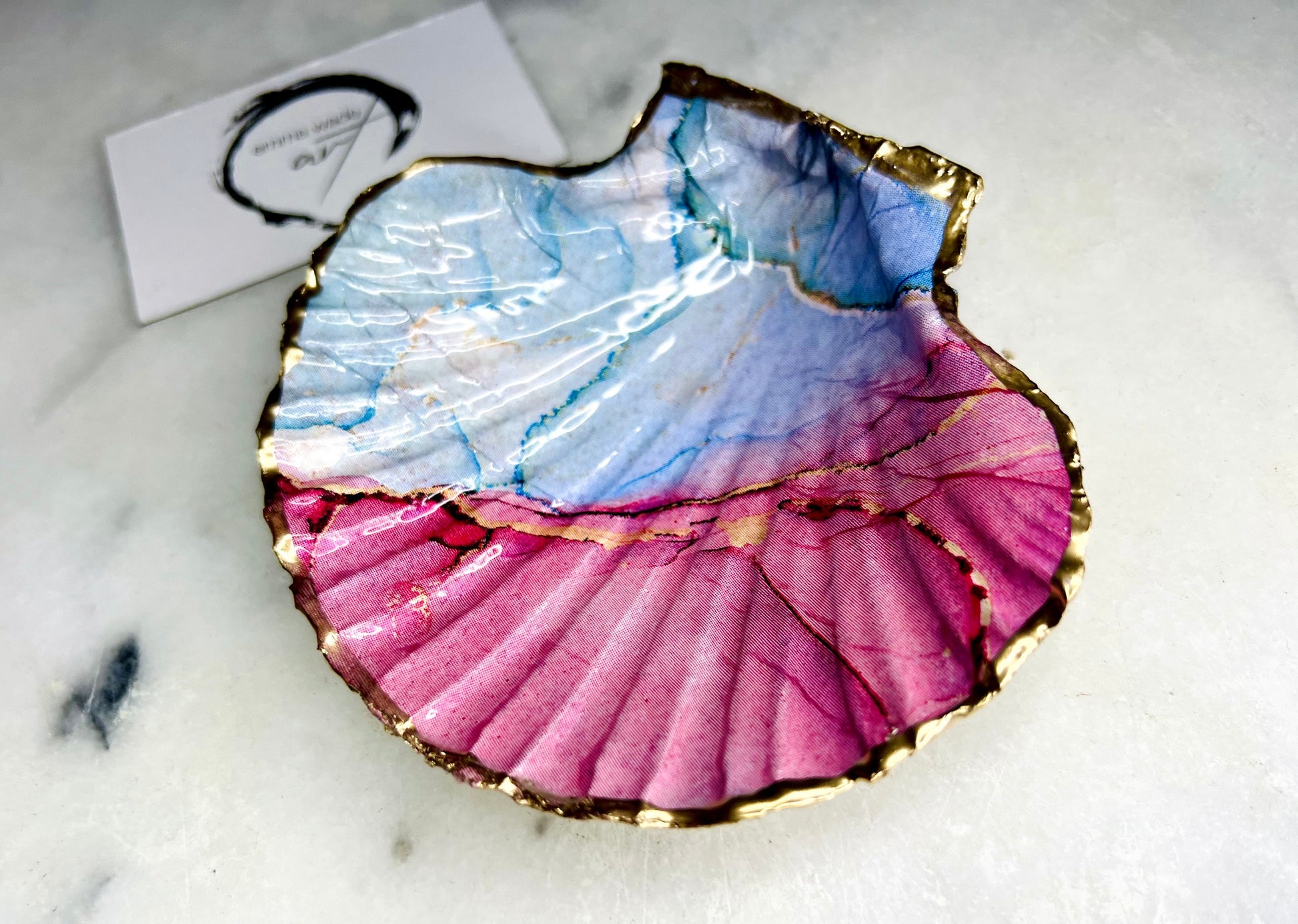A handmade scallop shell trinket dish painted in soft pink and pale blue tones in a flowing, watercolor-style pattern, finished with a gold leaf edge and sealed with a glossy clear resin. The natural shell shape is visible, with the ridged texture of the scallop shell showing through the design. The dish is shown on a light surface and works as a small decorative tray suitable for holding rings or jewellery in a bedroom or bathroom.