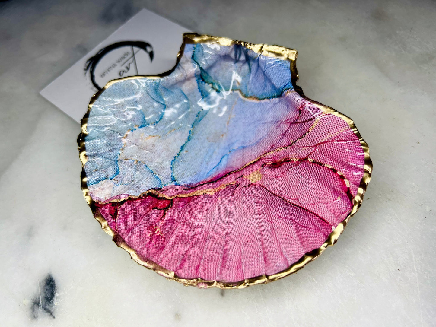 A handmade scallop shell trinket dish painted in soft pink and pale blue tones in a flowing, watercolor-style pattern, finished with a gold leaf edge and sealed with a glossy clear resin. The natural shell shape is visible, with the ridged texture of the scallop shell showing through the design. The dish is shown on a light surface and works as a small decorative tray suitable for holding rings or jewellery in a bedroom or bathroom.