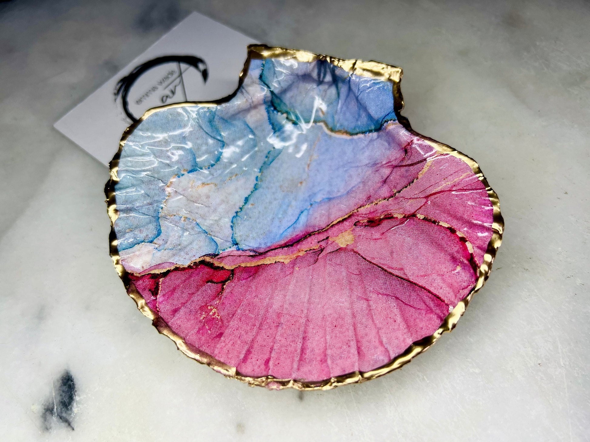A handmade scallop shell trinket dish painted in soft pink and pale blue tones in a flowing, watercolor-style pattern, finished with a gold leaf edge and sealed with a glossy clear resin. The natural shell shape is visible, with the ridged texture of the scallop shell showing through the design. The dish is shown on a light surface and works as a small decorative tray suitable for holding rings or jewellery in a bedroom or bathroom.