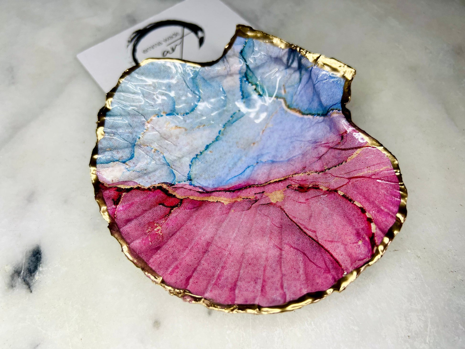 A handmade scallop shell trinket dish painted in soft pink and pale blue tones in a flowing, watercolor-style pattern, finished with a gold leaf edge and sealed with a glossy clear resin. The natural shell shape is visible, with the ridged texture of the scallop shell showing through the design. The dish is shown on a light surface and works as a small decorative tray suitable for holding rings or jewellery in a bedroom or bathroom.