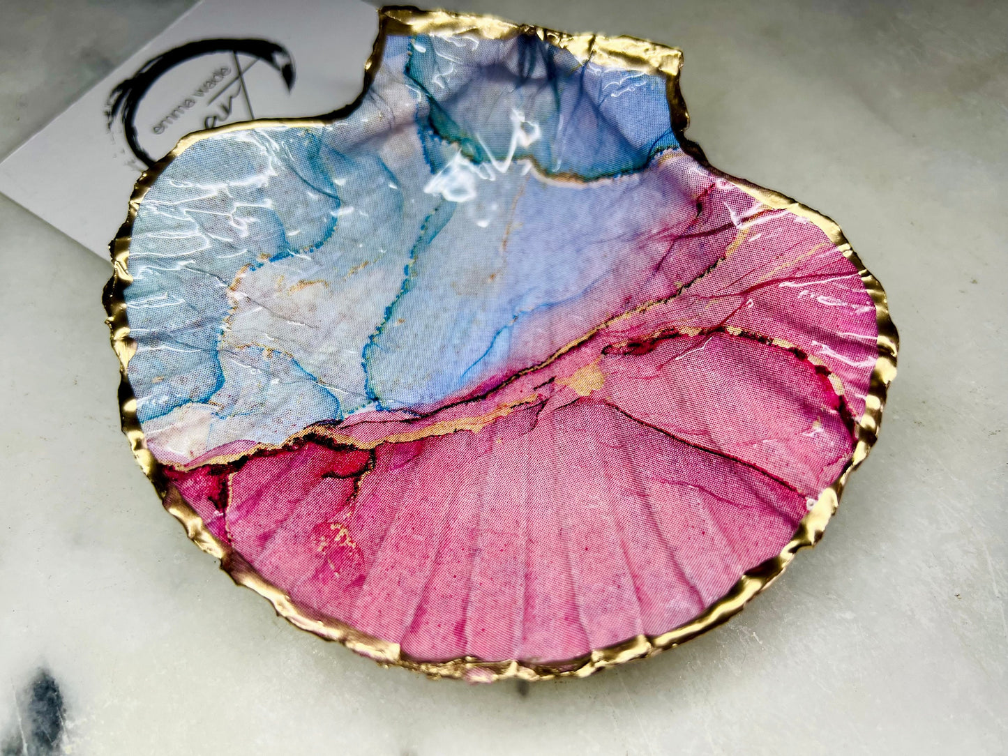 A handmade scallop shell trinket dish painted in soft pink and pale blue tones in a flowing, watercolor-style pattern, finished with a gold leaf edge and sealed with a glossy clear resin. The natural shell shape is visible, with the ridged texture of the scallop shell showing through the design. The dish is shown on a light surface and works as a small decorative tray suitable for holding rings or jewellery in a bedroom or bathroom.
