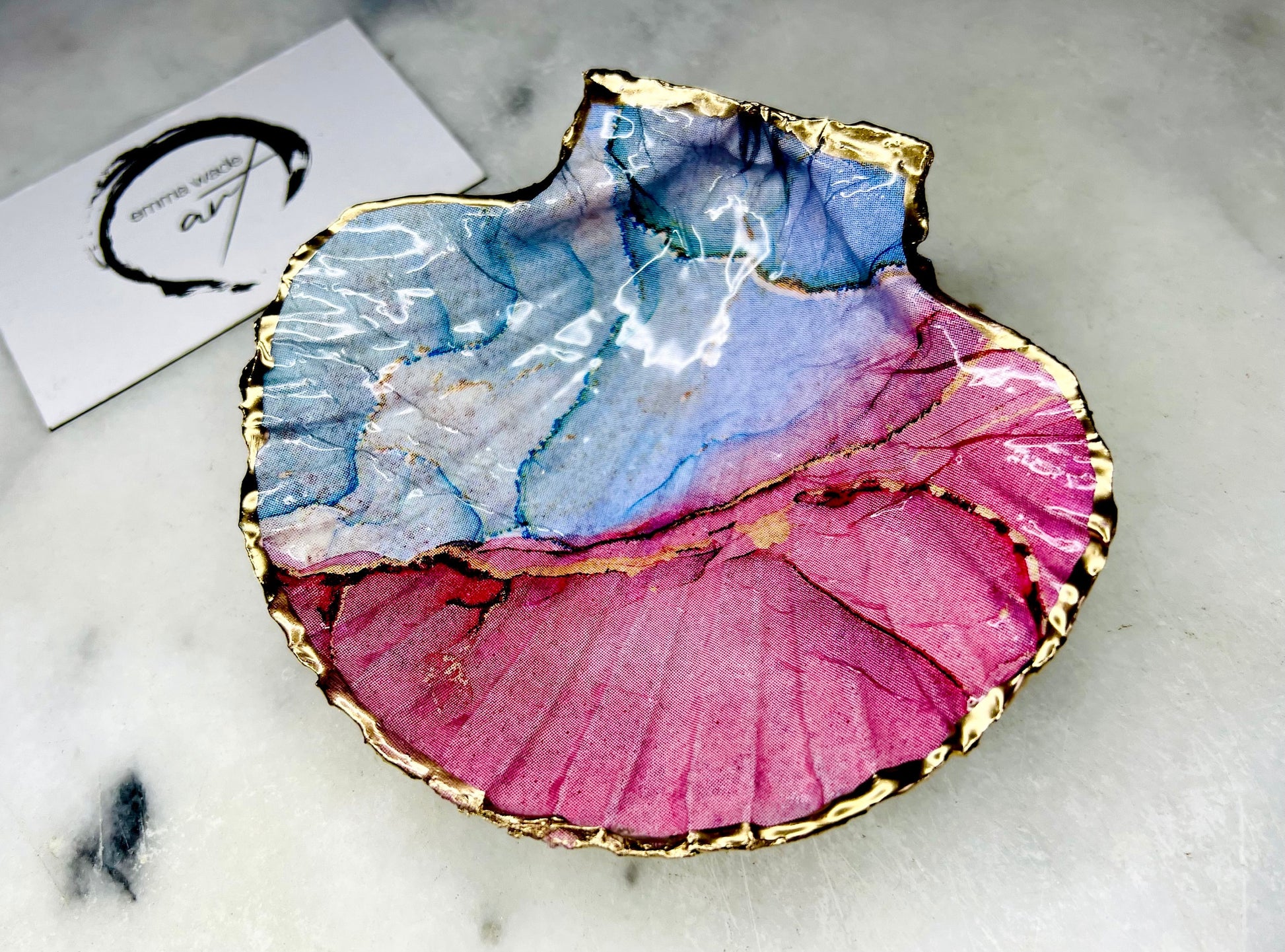 A handmade scallop shell trinket dish painted in soft pink and pale blue tones in a flowing, watercolor-style pattern, finished with a gold leaf edge and sealed with a glossy clear resin. The natural shell shape is visible, with the ridged texture of the scallop shell showing through the design. The dish is shown on a light surface and works as a small decorative tray suitable for holding rings or jewellery in a bedroom or bathroom.