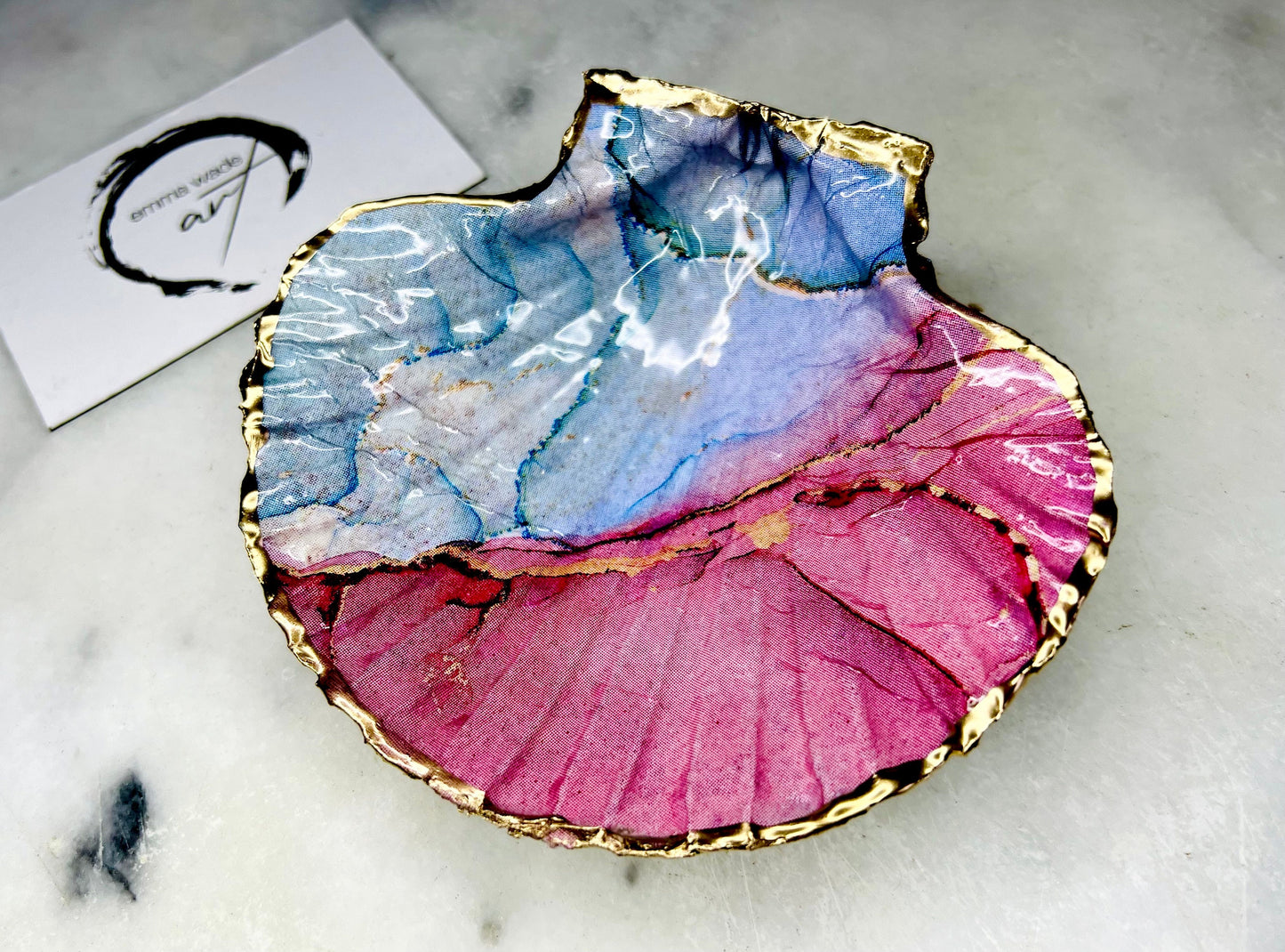 A handmade scallop shell trinket dish painted in soft pink and pale blue tones in a flowing, watercolor-style pattern, finished with a gold leaf edge and sealed with a glossy clear resin. The natural shell shape is visible, with the ridged texture of the scallop shell showing through the design. The dish is shown on a light surface and works as a small decorative tray suitable for holding rings or jewellery in a bedroom or bathroom.