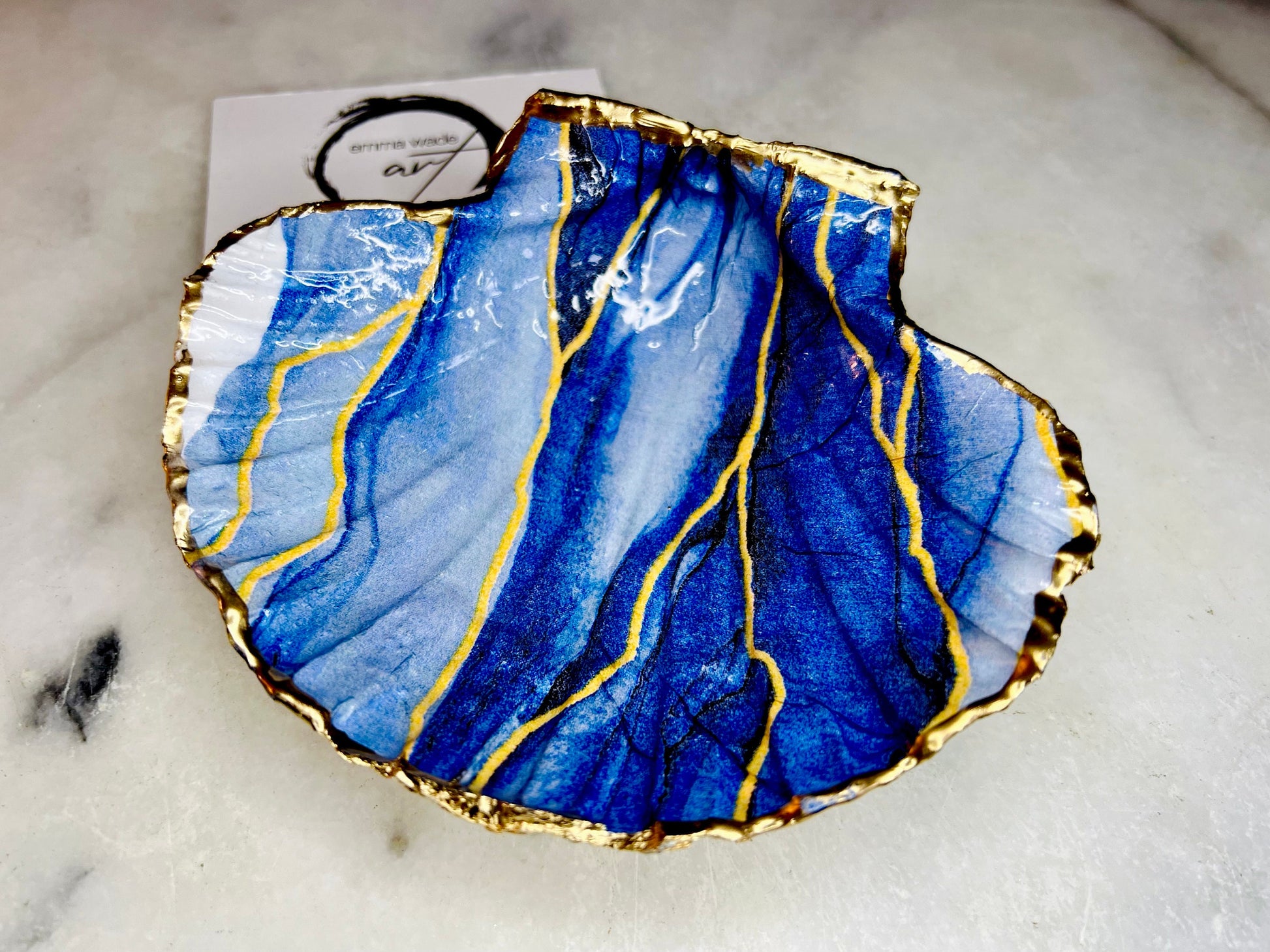 Hand painted blue waves scallop shell trinket dish with gold leaf accents and glossy resin finish, shown with optional gold driftwood display stand. Coastal jewellery and ring dish crafted from a natural Dorset scallop shell, featuring flowing blue wave artwork. Decorative shell bowl photographed on a light background, showing the natural shell shape and gilded edges, ideal for holding rings, earrings, trinkets or small treasures.