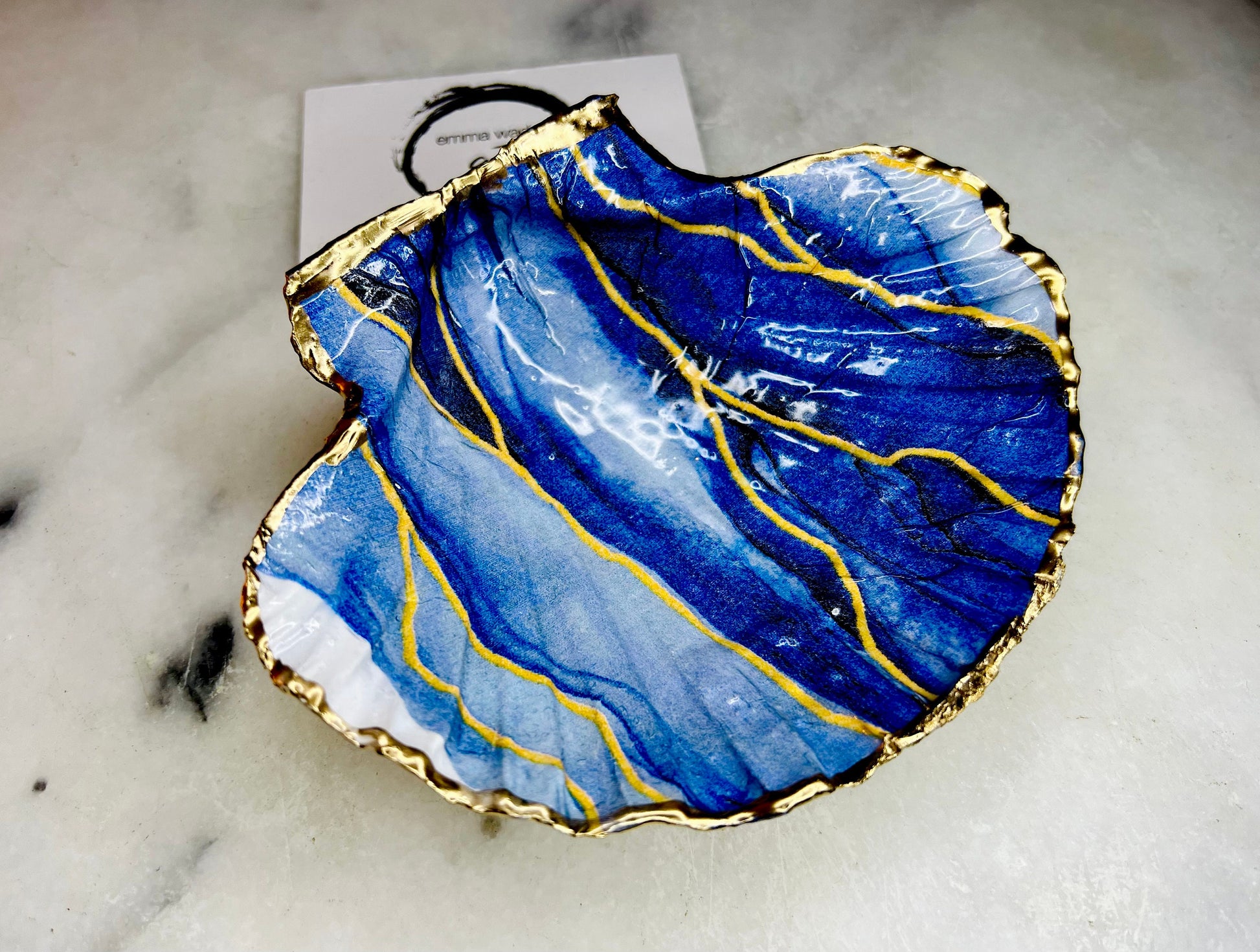 Hand painted blue waves scallop shell trinket dish with gold leaf accents and glossy resin finish, shown with optional gold driftwood display stand. Coastal jewellery and ring dish crafted from a natural Dorset scallop shell, featuring flowing blue wave artwork. Decorative shell bowl photographed on a light background, showing the natural shell shape and gilded edges, ideal for holding rings, earrings, trinkets or small treasures.