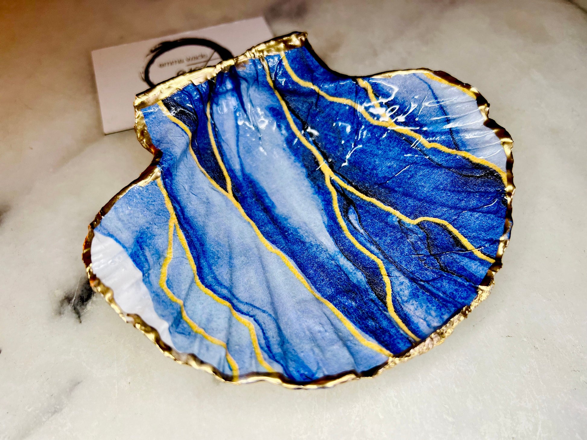 Hand painted blue waves scallop shell trinket dish with gold leaf accents and glossy resin finish, shown with optional gold driftwood display stand. Coastal jewellery and ring dish crafted from a natural Dorset scallop shell, featuring flowing blue wave artwork. Decorative shell bowl photographed on a light background, showing the natural shell shape and gilded edges, ideal for holding rings, earrings, trinkets or small treasures.