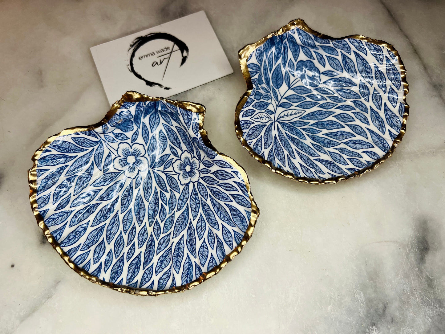 Handmade Dorset scallop shell trinket dishes featuring intricate blue floral and leaf artwork with hand-painted gold leaf edging and glossy crystal clear resin. Coastal botanical jewellery dishes inspired by Nordic Scandinavian decor, styled on a neutral background with elegant blue flower detailing and gilded edges.