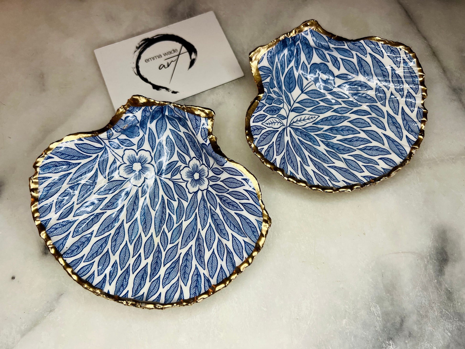 Handmade Dorset scallop shell trinket dishes featuring intricate blue floral and leaf artwork with hand-painted gold leaf edging and glossy crystal clear resin. Coastal botanical jewellery dishes inspired by Nordic Scandinavian decor, styled on a neutral background with elegant blue flower detailing and gilded edges.
