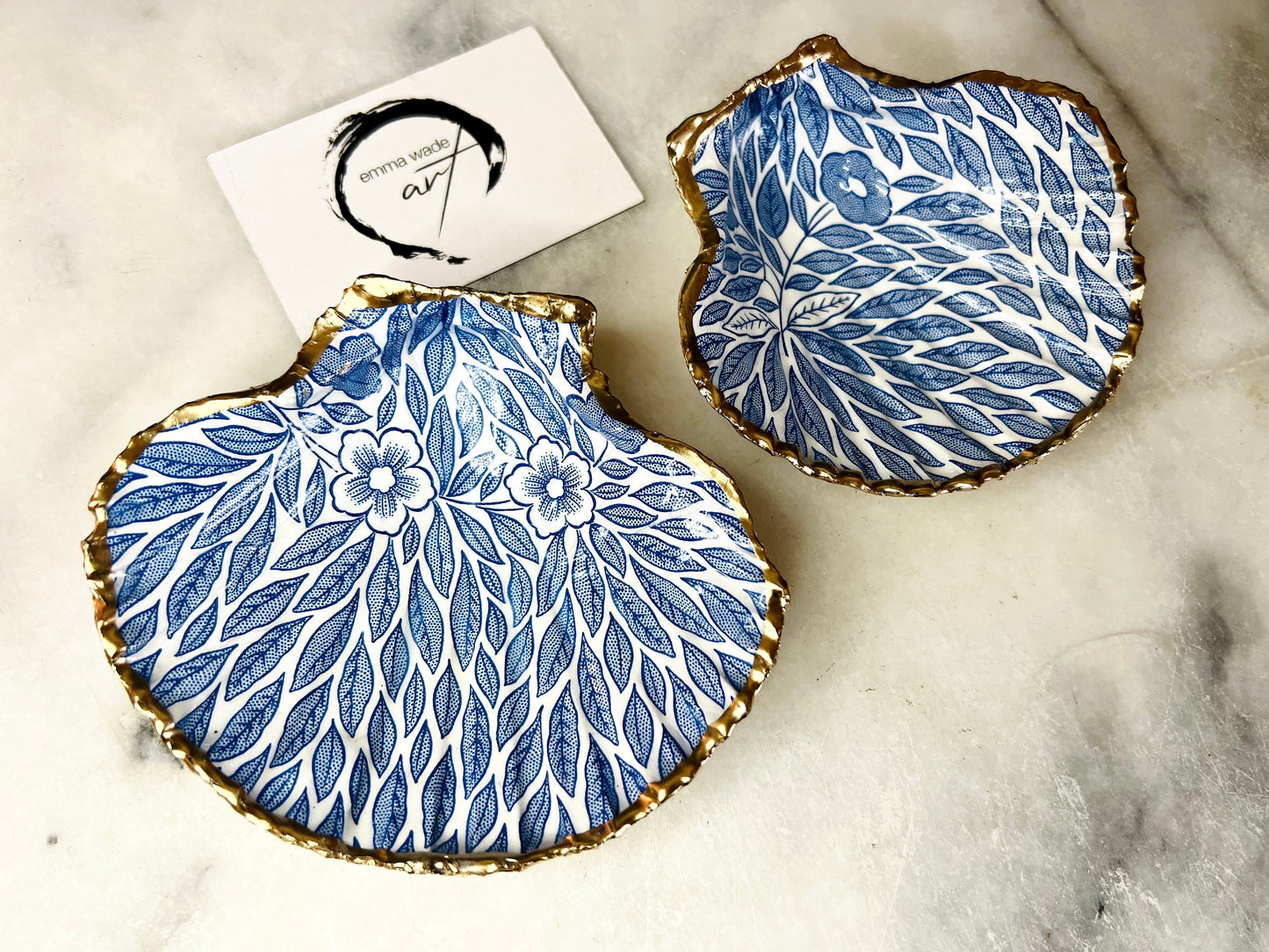 Handmade Dorset scallop shell trinket dishes featuring intricate blue floral and leaf artwork with hand-painted gold leaf edging and glossy crystal clear resin. Coastal botanical jewellery dishes inspired by Nordic Scandinavian decor, styled on a neutral background with elegant blue flower detailing and gilded edges.