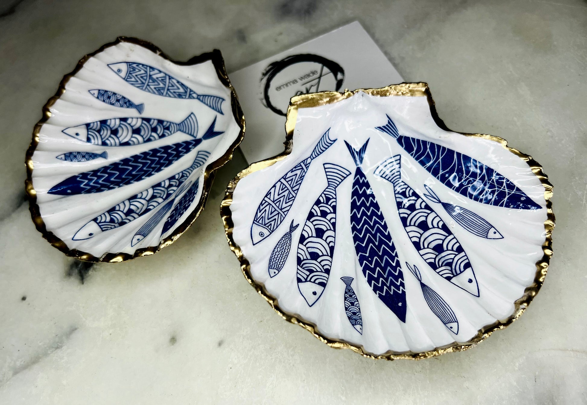 Handmade Dorset scallop shell trinket dishes featuring elegant blue fish illustrations with hand-painted gold leaf edging and glossy crystal clear resin. Coastal nautical jewellery dishes inspired by Scandinavian ocean decor, styled on a neutral background with detailed blue fish artwork and luxurious gilded edges.