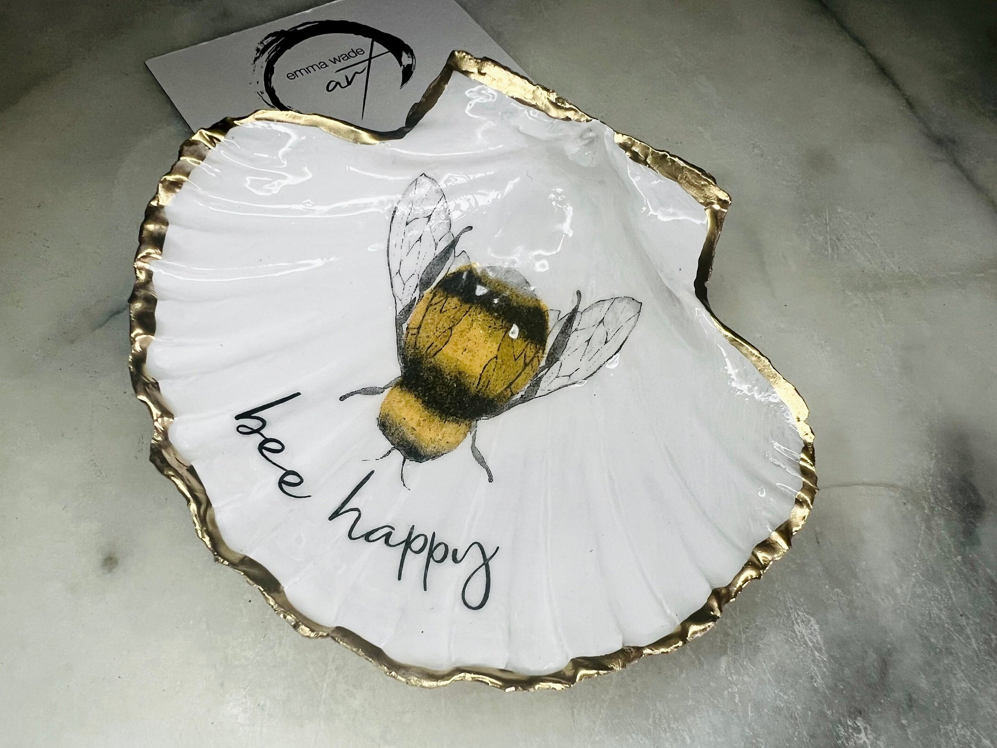 Handmade scallop shell trinket dish featuring a bumblebee illustration with gold leaf edging and glossy resin finish. Coastal jewellery dish in natural white shell with bee artwork, displayed as a ring holder and handmade bee lover gift