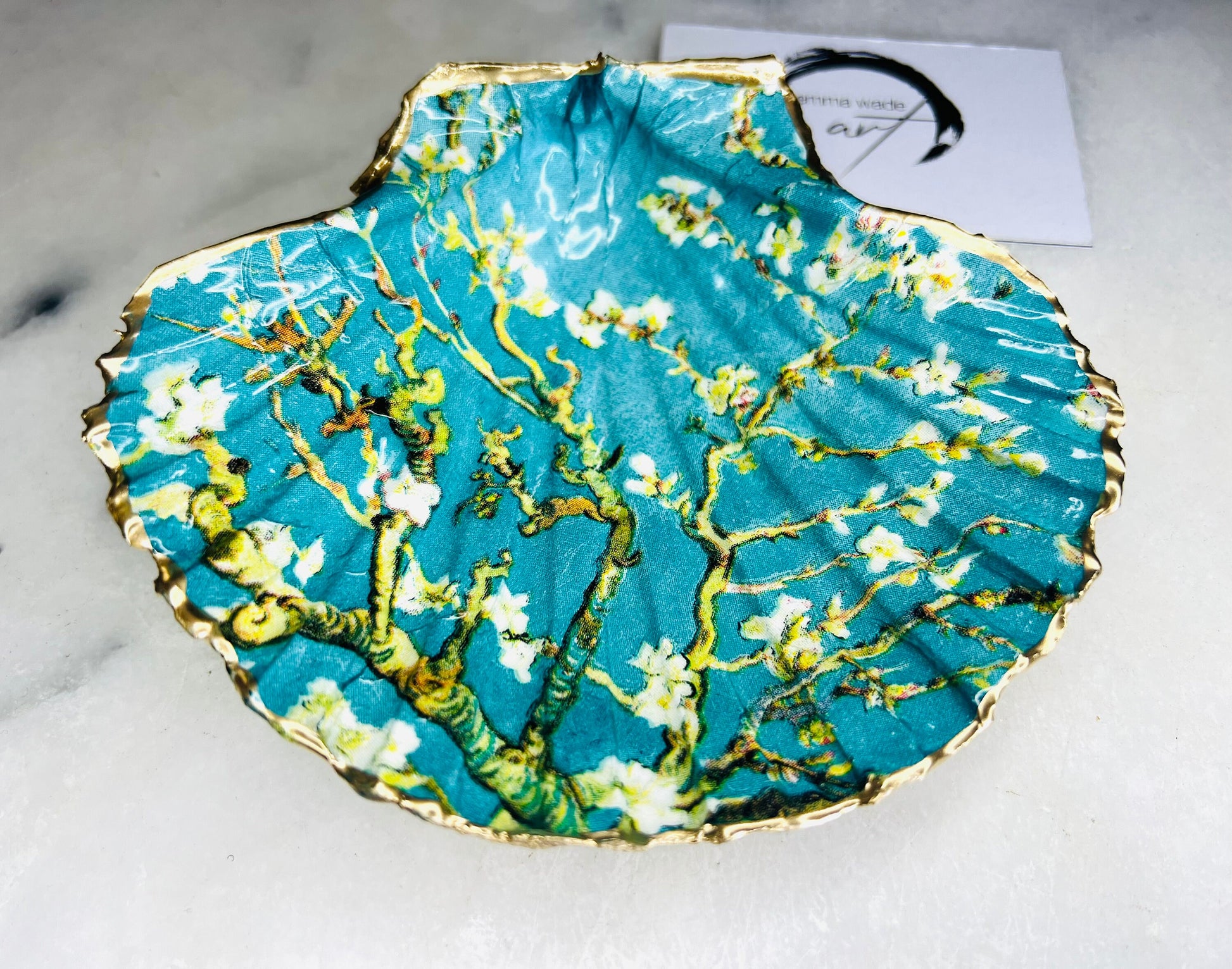 Handcrafted Dorset scallop shell trinket dish inspired by Vincent van Gogh’s Almond Blossom painting. Turquoise blue and green background with branching blossom design and raised gold detailing, finished with liquid gold leaf edging and sealed in high-gloss resin. Decorative shell jewellery holder styled on marble surface, suitable for rings, earrings, trinkets or as a tea light holder. Coastal art-inspired home decor piece.