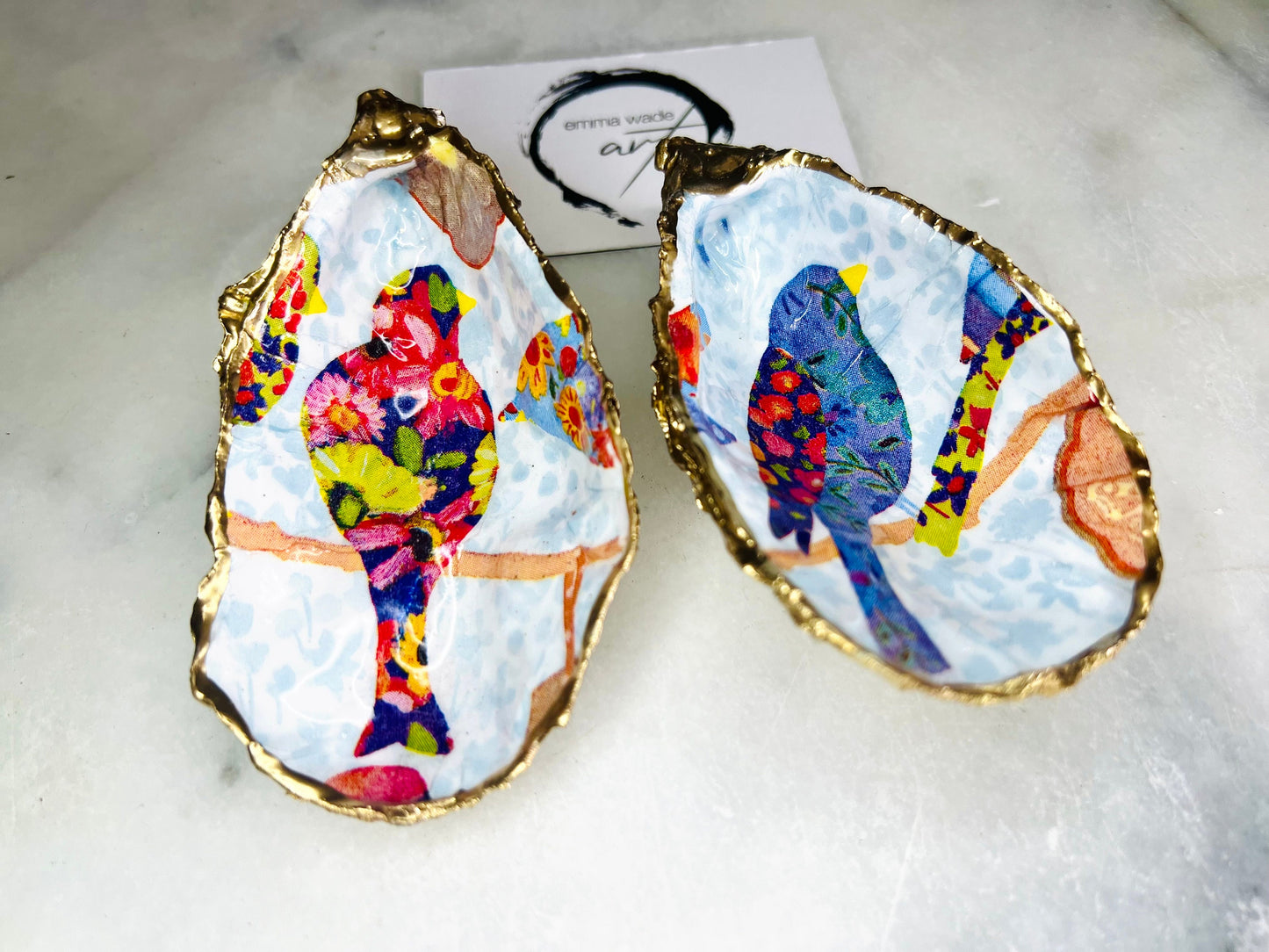 Handmade Dorset oyster shell trinket dishes featuring colourful bird artwork in pink and blue tones, finished with hand-painted gold leaf edging and glossy resin. Decorative coastal jewellery dishes styled on a white marble background with vibrant nature-inspired bird illustrations.