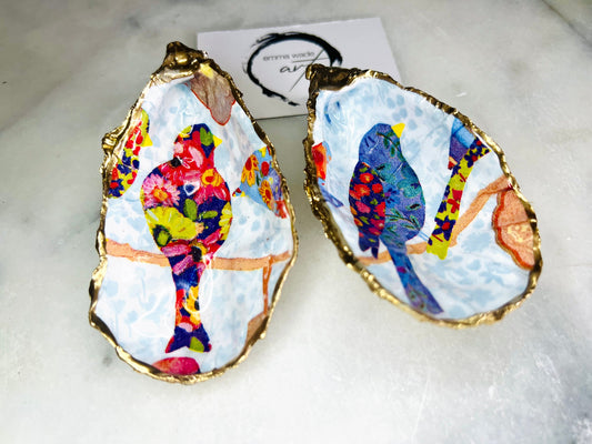 Handmade Dorset oyster shell trinket dishes featuring colourful bird artwork in pink and blue tones, finished with hand-painted gold leaf edging and glossy resin. Decorative coastal jewellery dishes styled on a white marble background with vibrant nature-inspired bird illustrations.