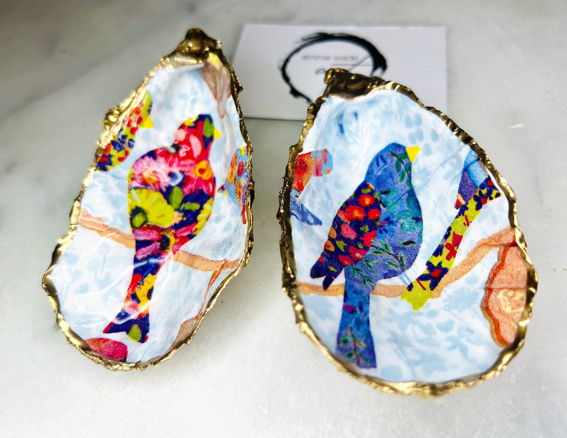 Handmade Dorset oyster shell trinket dishes featuring colourful bird artwork in pink and blue tones, finished with hand-painted gold leaf edging and glossy resin. Decorative coastal jewellery dishes styled on a white marble background with vibrant nature-inspired bird illustrations.