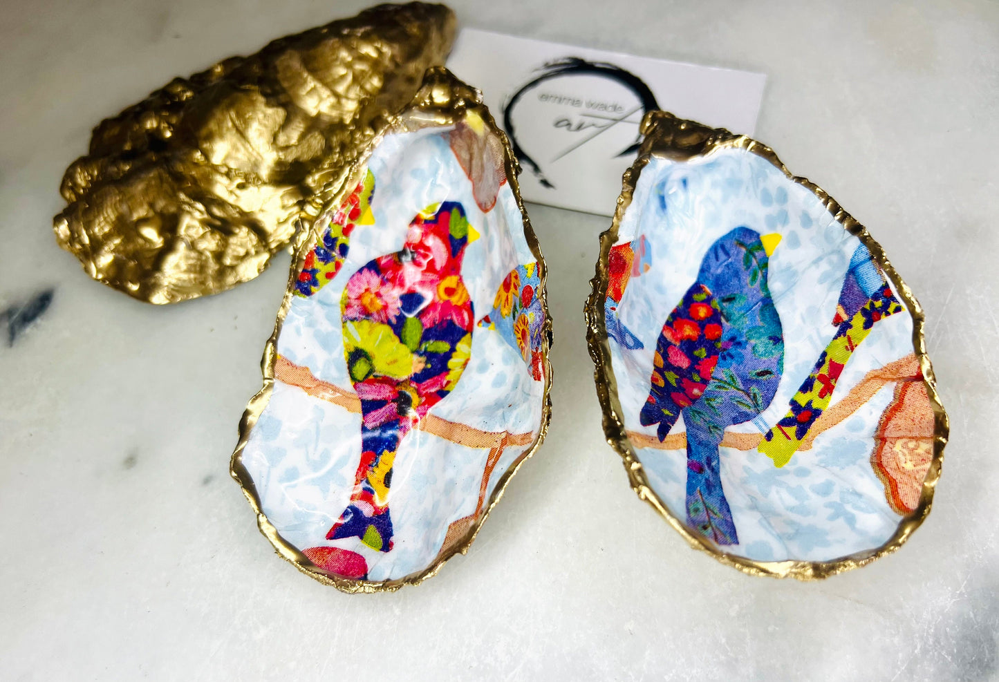 Handmade Dorset oyster shell trinket dishes featuring colourful bird artwork in pink and blue tones, finished with hand-painted gold leaf edging and glossy resin. Decorative coastal jewellery dishes styled on a white marble background with vibrant nature-inspired bird illustrations.