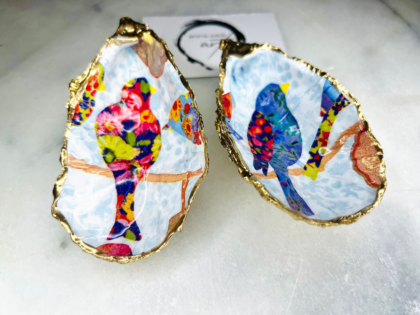Handmade Dorset oyster shell trinket dishes featuring colourful bird artwork in pink and blue tones, finished with hand-painted gold leaf edging and glossy resin. Decorative coastal jewellery dishes styled on a white marble background with vibrant nature-inspired bird illustrations.