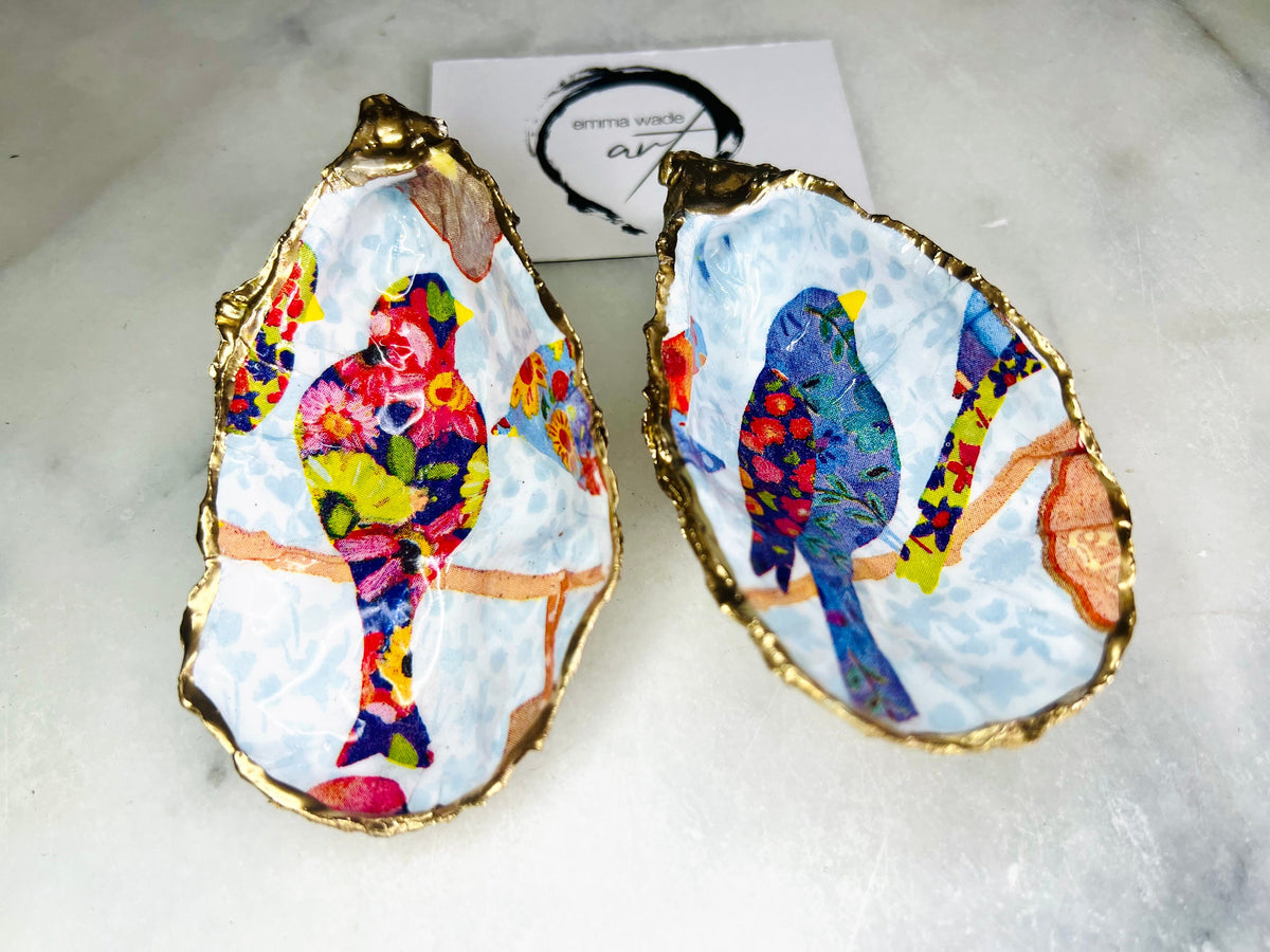 Handmade Dorset oyster shell trinket dishes featuring colourful bird artwork in pink and blue tones, finished with hand-painted gold leaf edging and glossy resin. Decorative coastal jewellery dishes styled on a white marble background with vibrant nature-inspired bird illustrations.