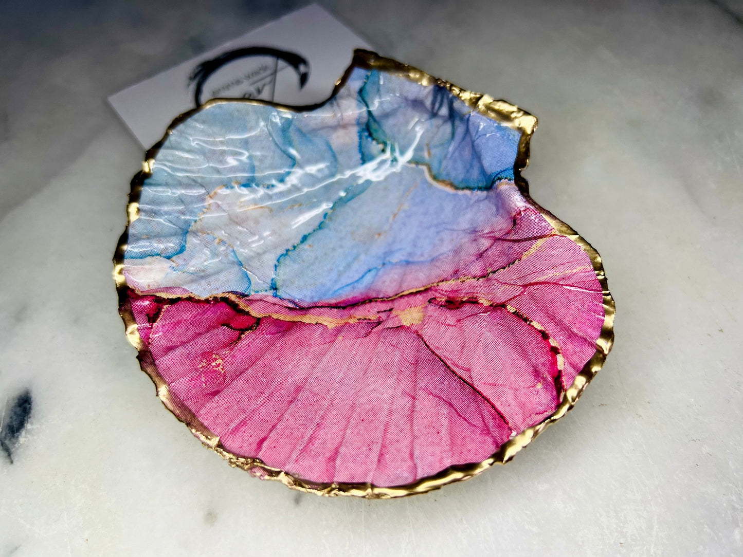 A handmade scallop shell trinket dish painted in soft pink and pale blue tones in a flowing, watercolor-style pattern, finished with a gold leaf edge and sealed with a glossy clear resin. The natural shell shape is visible, with the ridged texture of the scallop shell showing through the design. The dish is shown on a light surface and works as a small decorative tray suitable for holding rings or jewellery in a bedroom or bathroom.