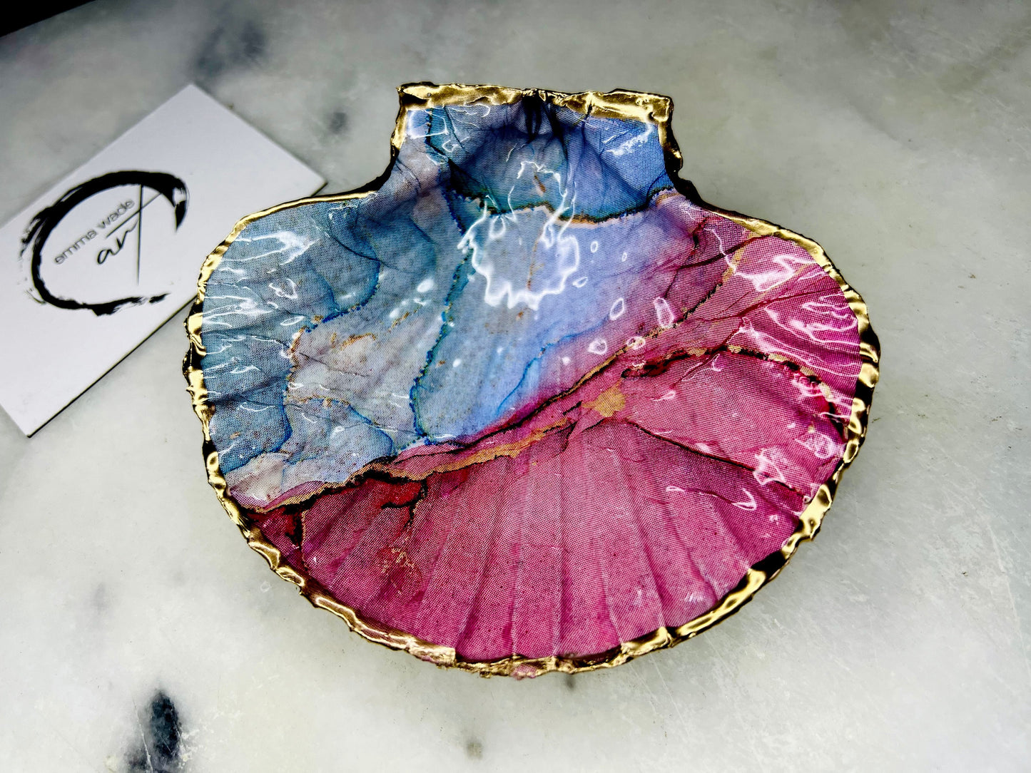 A handmade scallop shell trinket dish painted in soft pink and pale blue tones in a flowing, watercolor-style pattern, finished with a gold leaf edge and sealed with a glossy clear resin. The natural shell shape is visible, with the ridged texture of the scallop shell showing through the design. The dish is shown on a light surface and works as a small decorative tray suitable for holding rings or jewellery in a bedroom or bathroom.