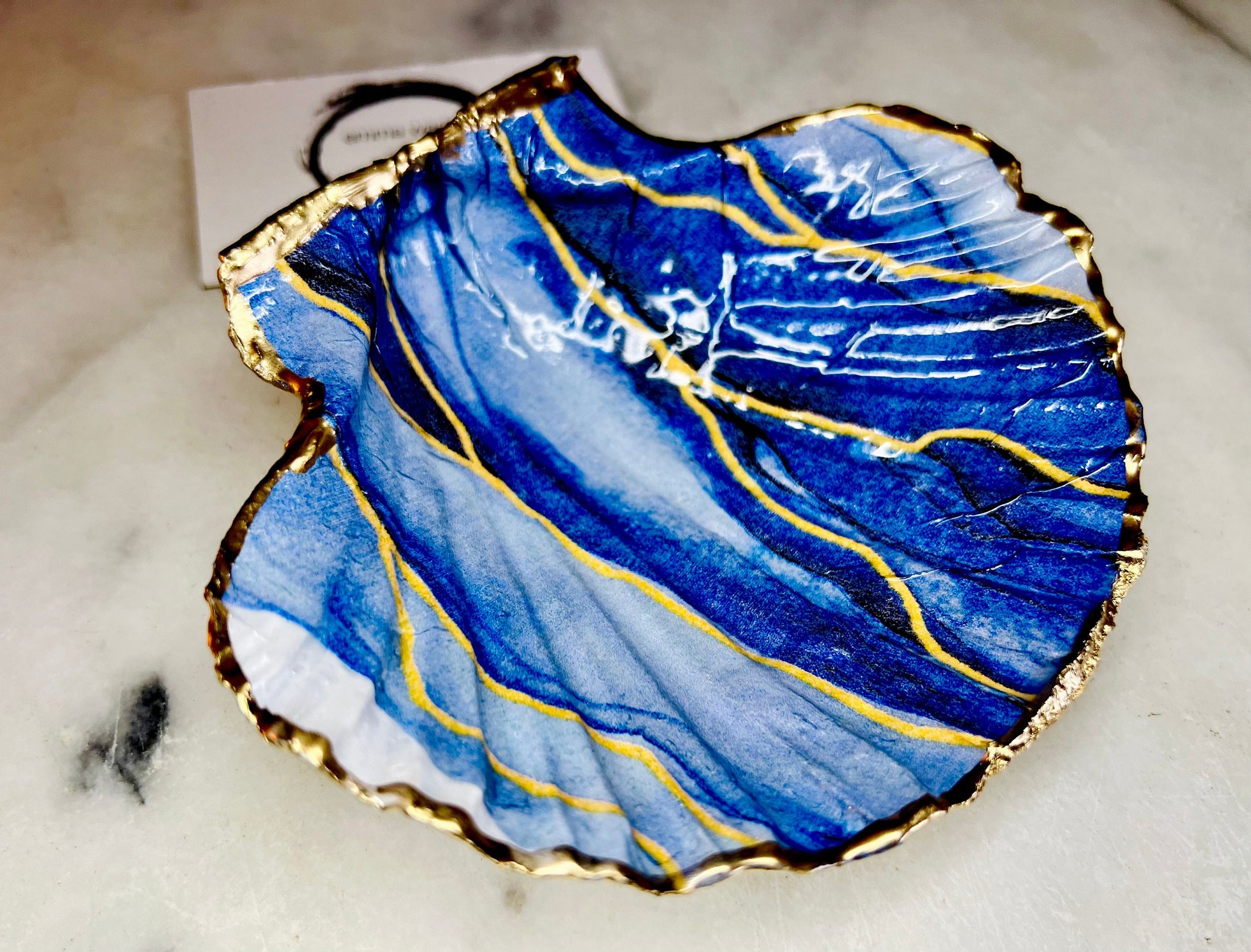 Hand painted blue waves scallop shell trinket dish with gold leaf accents and glossy resin finish, shown with optional gold driftwood display stand. Coastal jewellery and ring dish crafted from a natural Dorset scallop shell, featuring flowing blue wave artwork. Decorative shell bowl photographed on a light background, showing the natural shell shape and gilded edges, ideal for holding rings, earrings, trinkets or small treasures.