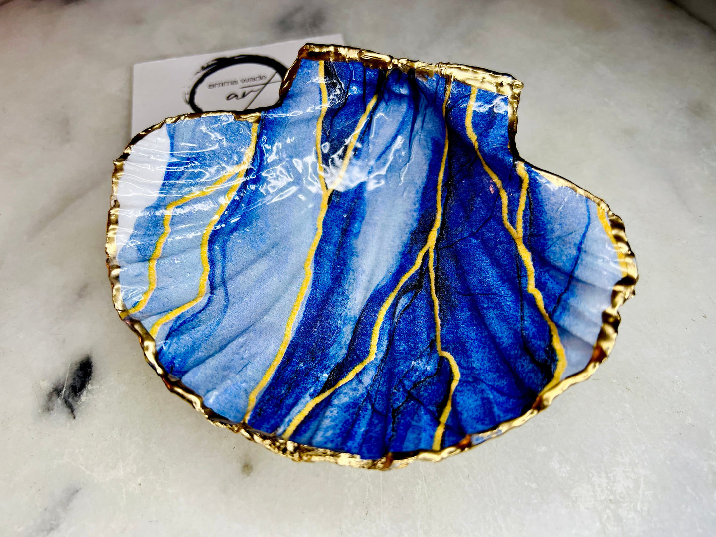 Hand painted blue waves scallop shell trinket dish with gold leaf accents and glossy resin finish, shown with optional gold driftwood display stand. Coastal jewellery and ring dish crafted from a natural Dorset scallop shell, featuring flowing blue wave artwork. Decorative shell bowl photographed on a light background, showing the natural shell shape and gilded edges, ideal for holding rings, earrings, trinkets or small treasures.