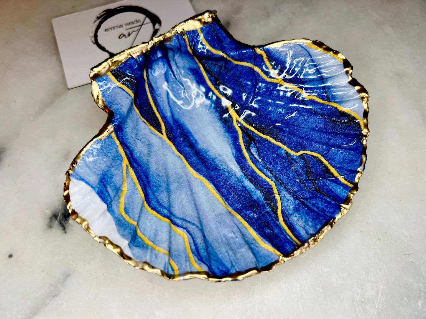 Hand painted blue waves scallop shell trinket dish with gold leaf accents and glossy resin finish, shown with optional gold driftwood display stand. Coastal jewellery and ring dish crafted from a natural Dorset scallop shell, featuring flowing blue wave artwork. Decorative shell bowl photographed on a light background, showing the natural shell shape and gilded edges, ideal for holding rings, earrings, trinkets or small treasures.
