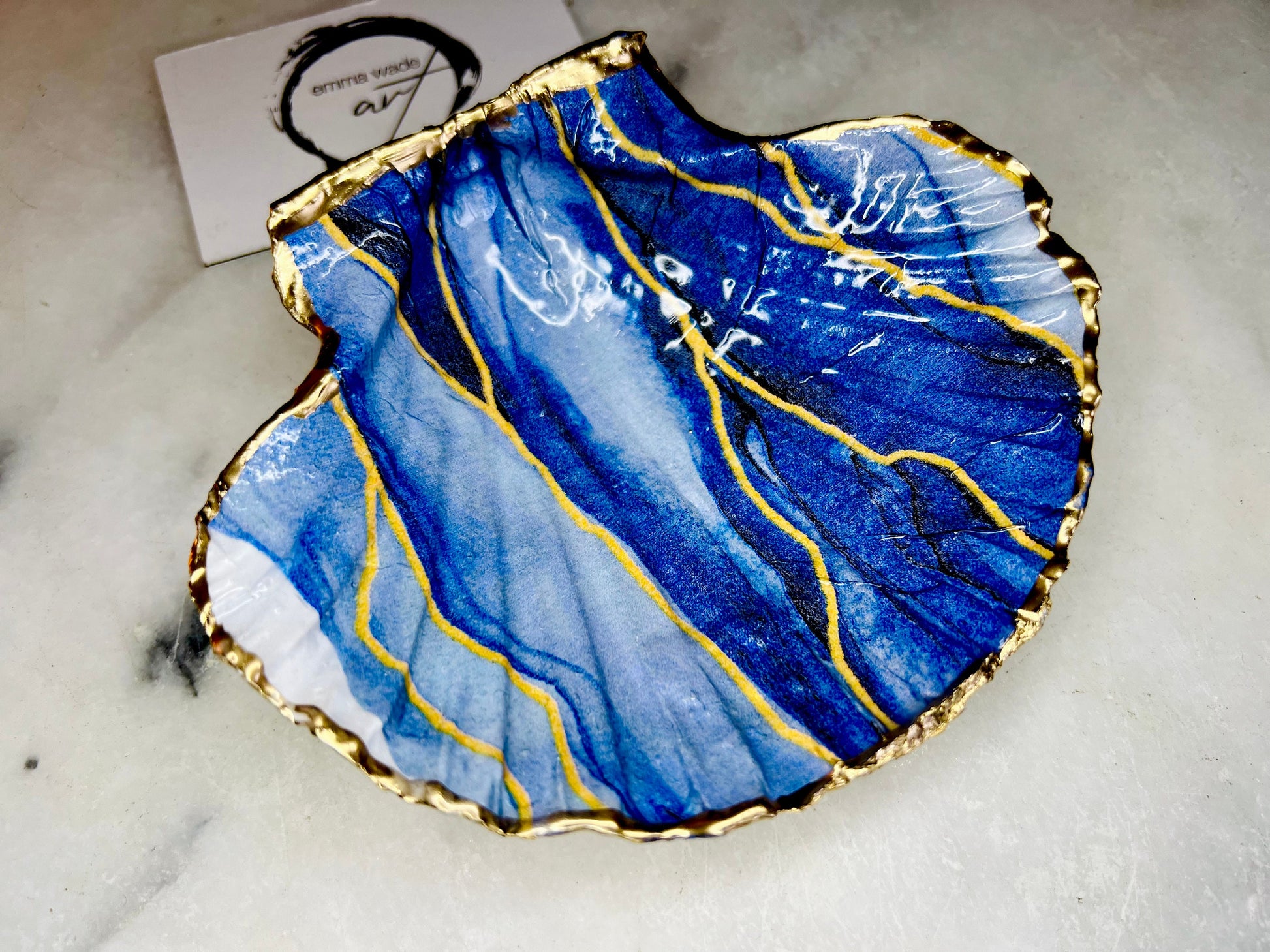 Hand painted blue waves scallop shell trinket dish with gold leaf accents and glossy resin finish, shown with optional gold driftwood display stand. Coastal jewellery and ring dish crafted from a natural Dorset scallop shell, featuring flowing blue wave artwork. Decorative shell bowl photographed on a light background, showing the natural shell shape and gilded edges, ideal for holding rings, earrings, trinkets or small treasures.