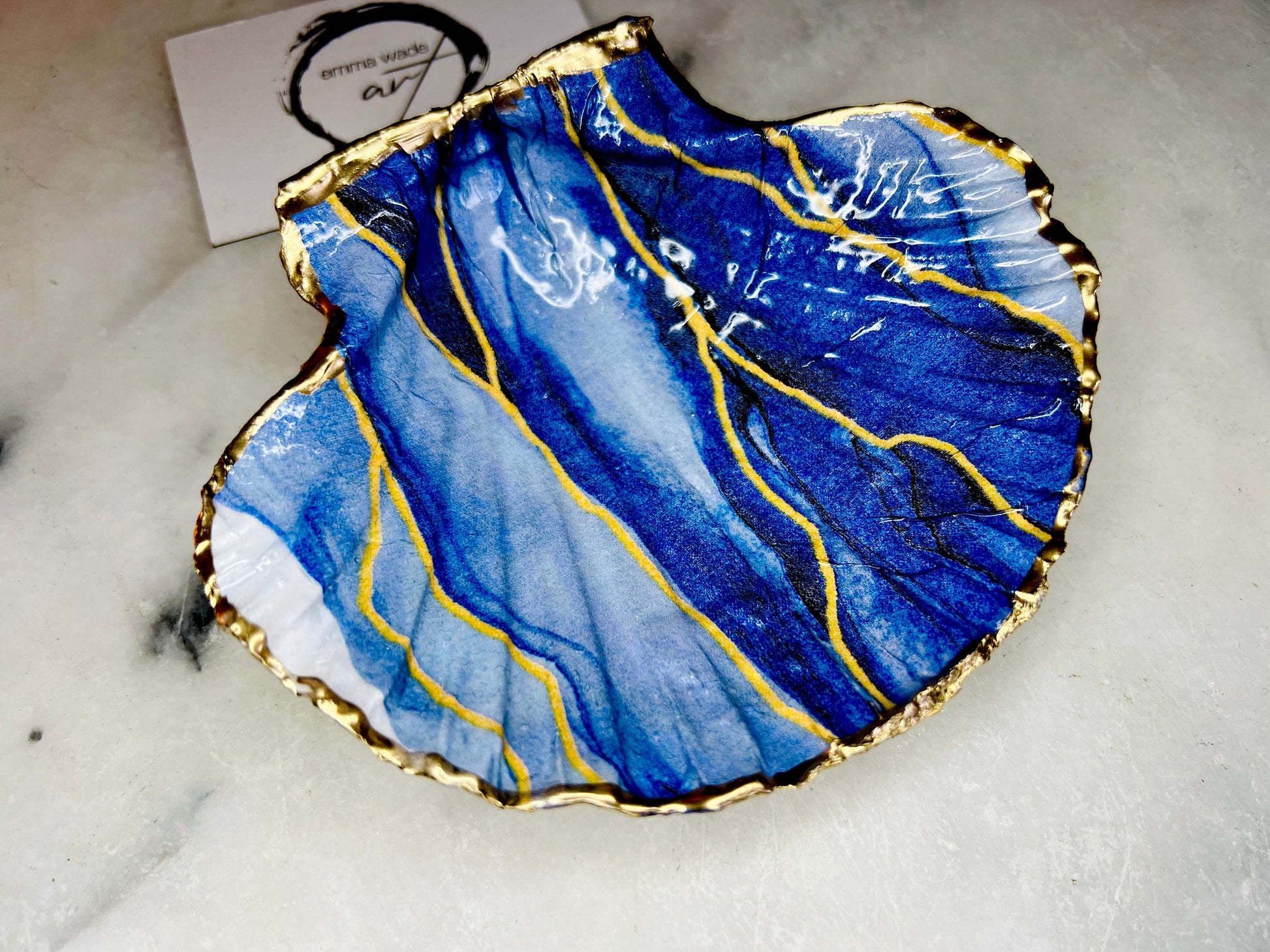 Hand painted blue waves scallop shell trinket dish with gold leaf accents and glossy resin finish, shown with optional gold driftwood display stand. Coastal jewellery and ring dish crafted from a natural Dorset scallop shell, featuring flowing blue wave artwork. Decorative shell bowl photographed on a light background, showing the natural shell shape and gilded edges, ideal for holding rings, earrings, trinkets or small treasures.
