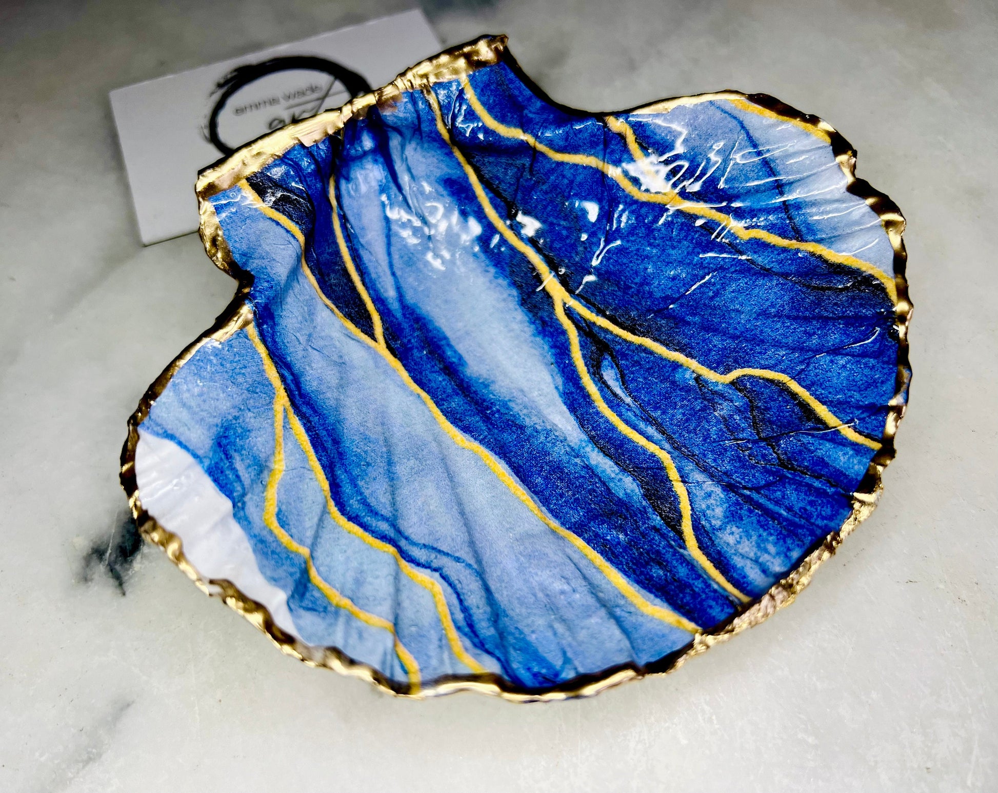 Hand painted blue waves scallop shell trinket dish with gold leaf accents and glossy resin finish, shown with optional gold driftwood display stand. Coastal jewellery and ring dish crafted from a natural Dorset scallop shell, featuring flowing blue wave artwork. Decorative shell bowl photographed on a light background, showing the natural shell shape and gilded edges, ideal for holding rings, earrings, trinkets or small treasures.