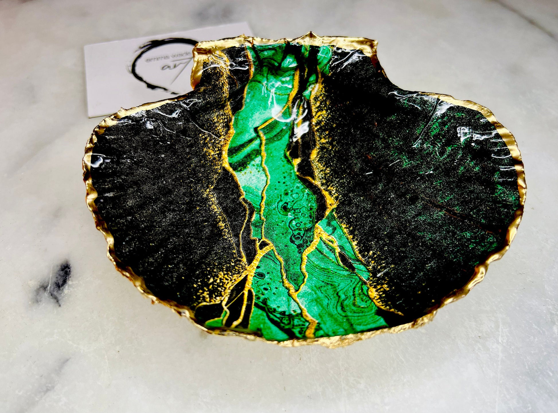 A handmade scallop shell trinket dish painted in deep green and black tones with an agate-style pattern and finished with a gold leaf edge, sealed with a glossy clear resin. The natural ridges and shape of the scallop shell are visible beneath the design, giving the piece texture and depth. The dish is shown as a small decorative tray suitable for holding rings or jewellery and works as a coastal-inspired accent in a bedroom, bathroom, or dressing area.