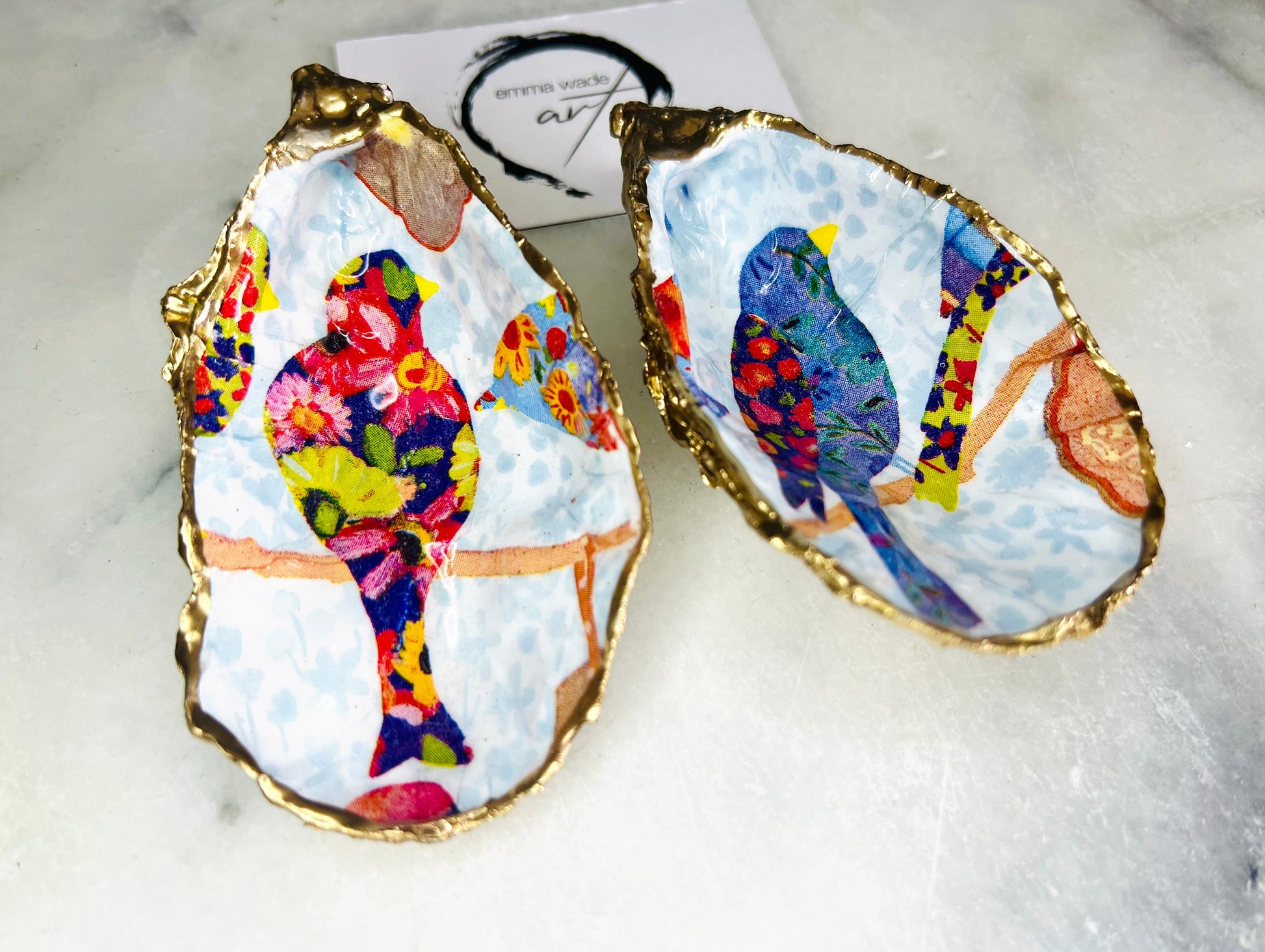 Handmade Dorset oyster shell trinket dishes featuring colourful bird artwork in pink and blue tones, finished with hand-painted gold leaf edging and glossy resin. Decorative coastal jewellery dishes styled on a white marble background with vibrant nature-inspired bird illustrations.