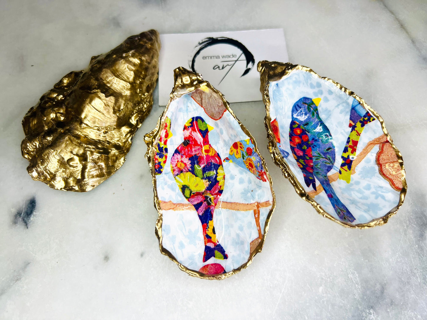 Handmade Dorset oyster shell trinket dishes featuring colourful bird artwork in pink and blue tones, finished with hand-painted gold leaf edging and glossy resin. Decorative coastal jewellery dishes styled on a white marble background with vibrant nature-inspired bird illustrations.