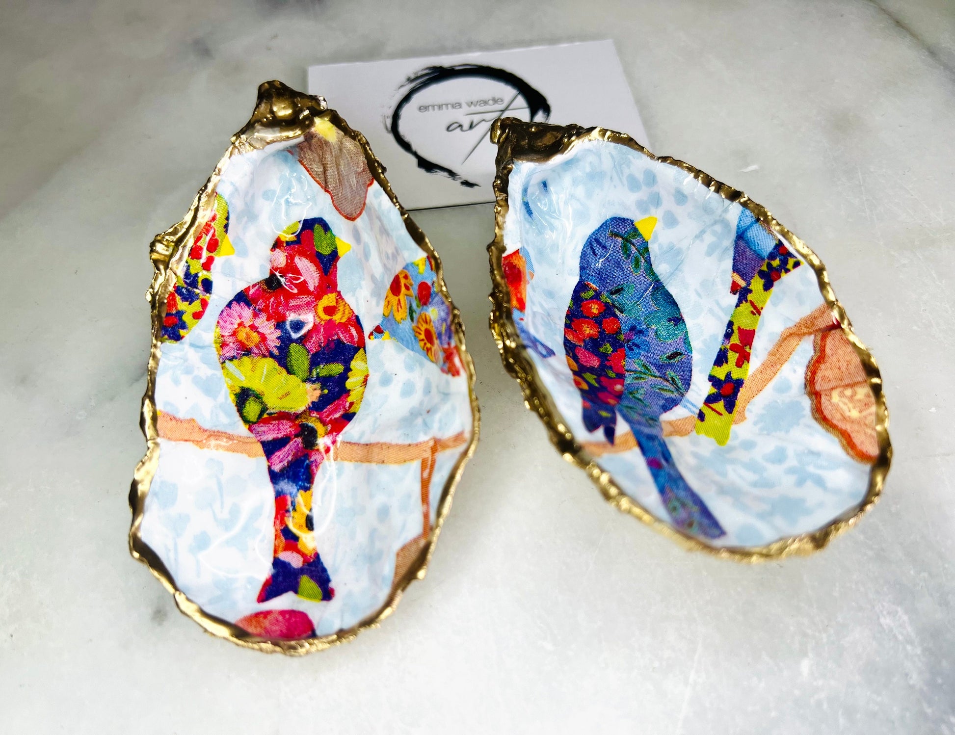 Handmade Dorset oyster shell trinket dishes featuring colourful bird artwork in pink and blue tones, finished with hand-painted gold leaf edging and glossy resin. Decorative coastal jewellery dishes styled on a white marble background with vibrant nature-inspired bird illustrations.