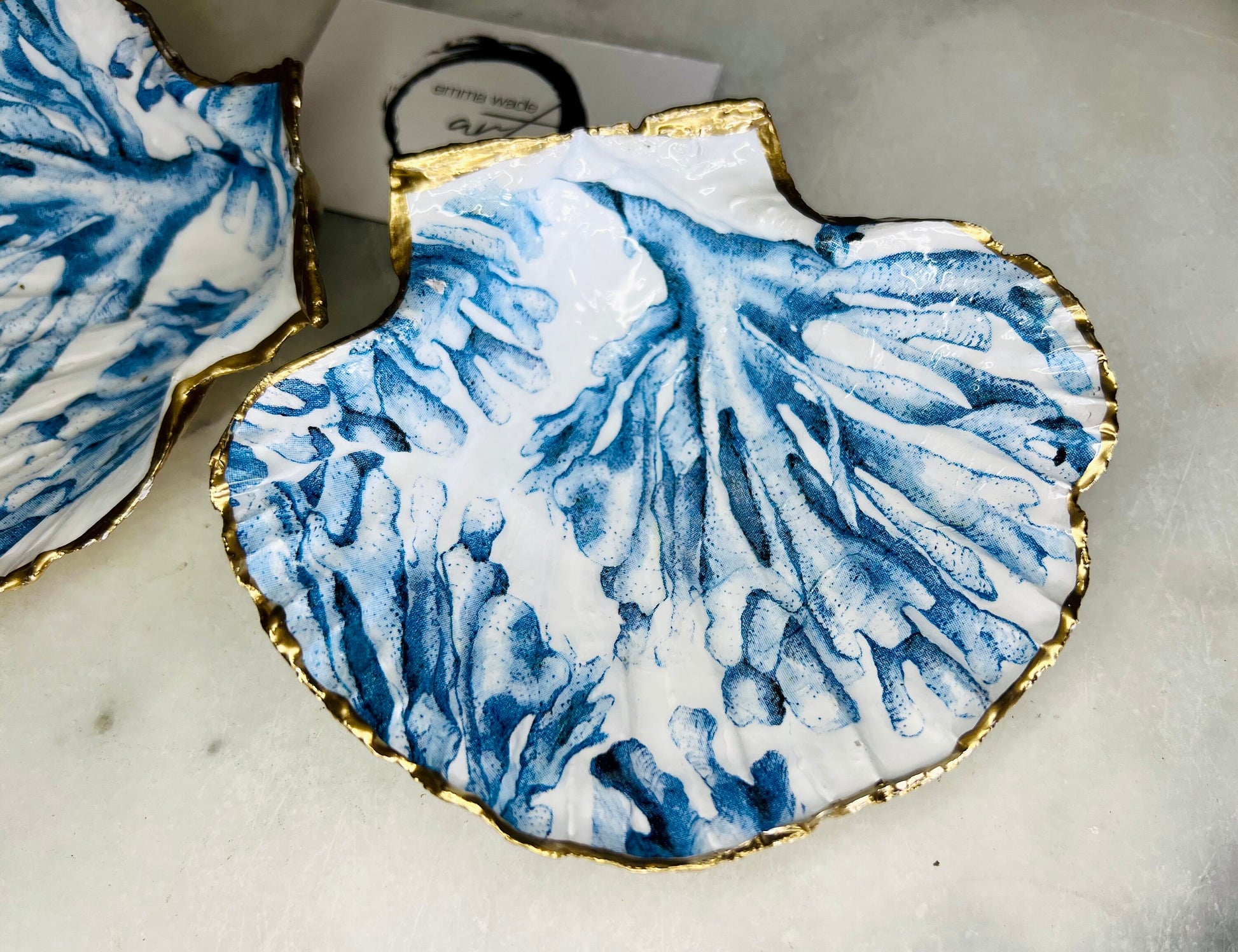 Blue seaweed scallop shell ring catcher with hand applied gold leaf edging and glossy resin finish. Handmade Dorset shell jewellery tray with ocean inspired design styled on marble background. Coastal vanity decor and beach house accessory with optional gold driftwood stand.