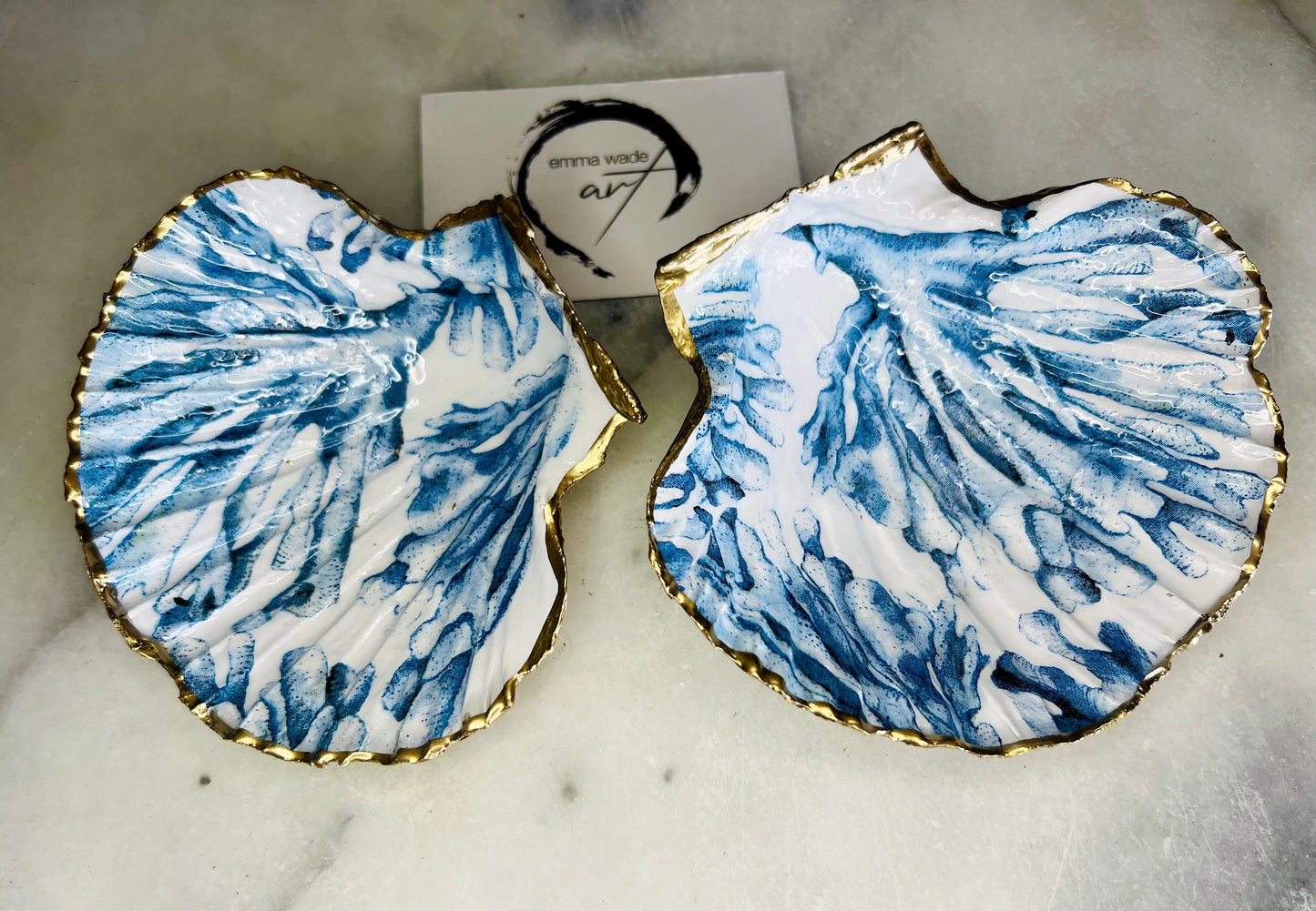 Blue seaweed scallop shell ring catcher with hand applied gold leaf edging and glossy resin finish. Handmade Dorset shell jewellery tray with ocean inspired design styled on marble background. Coastal vanity decor and beach house accessory with optional gold driftwood stand.