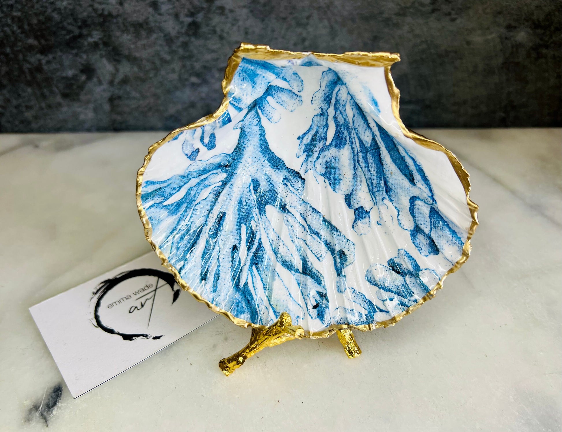 Blue seaweed scallop shell ring catcher with hand applied gold leaf edging and glossy resin finish. Handmade Dorset shell jewellery tray with ocean inspired design styled on marble background. Coastal vanity decor and beach house accessory with optional gold driftwood stand.