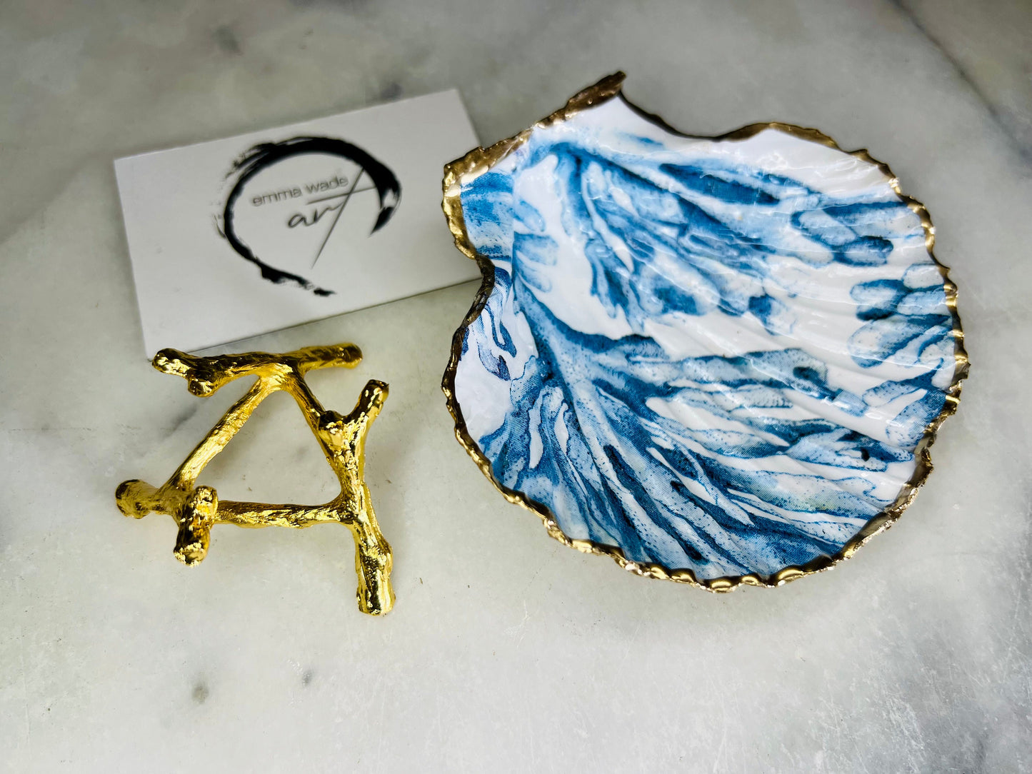 Blue seaweed scallop shell ring catcher with hand applied gold leaf edging and glossy resin finish. Handmade Dorset shell jewellery tray with ocean inspired design styled on marble background. Coastal vanity decor and beach house accessory with optional gold driftwood stand.