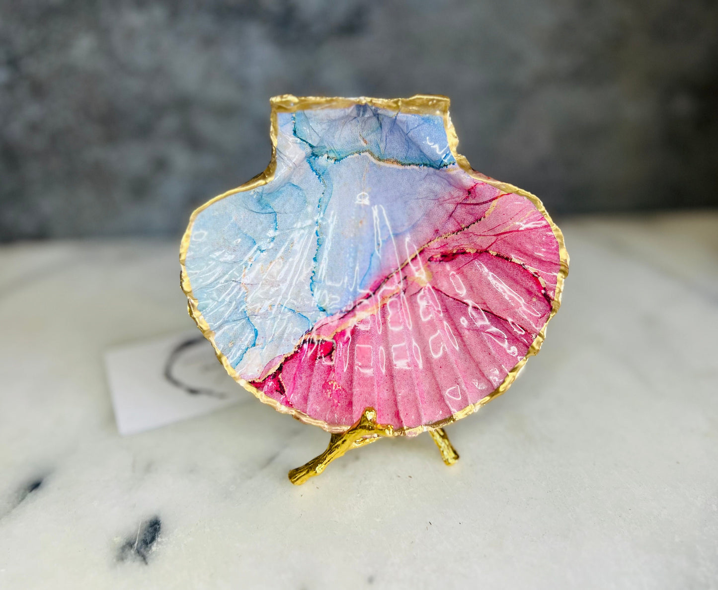 A handmade scallop shell trinket dish painted in soft pink and pale blue tones in a flowing, watercolor-style pattern, finished with a gold leaf edge and sealed with a glossy clear resin. The natural shell shape is visible, with the ridged texture of the scallop shell showing through the design. The dish is shown on a light surface and works as a small decorative tray suitable for holding rings or jewellery in a bedroom or bathroom.