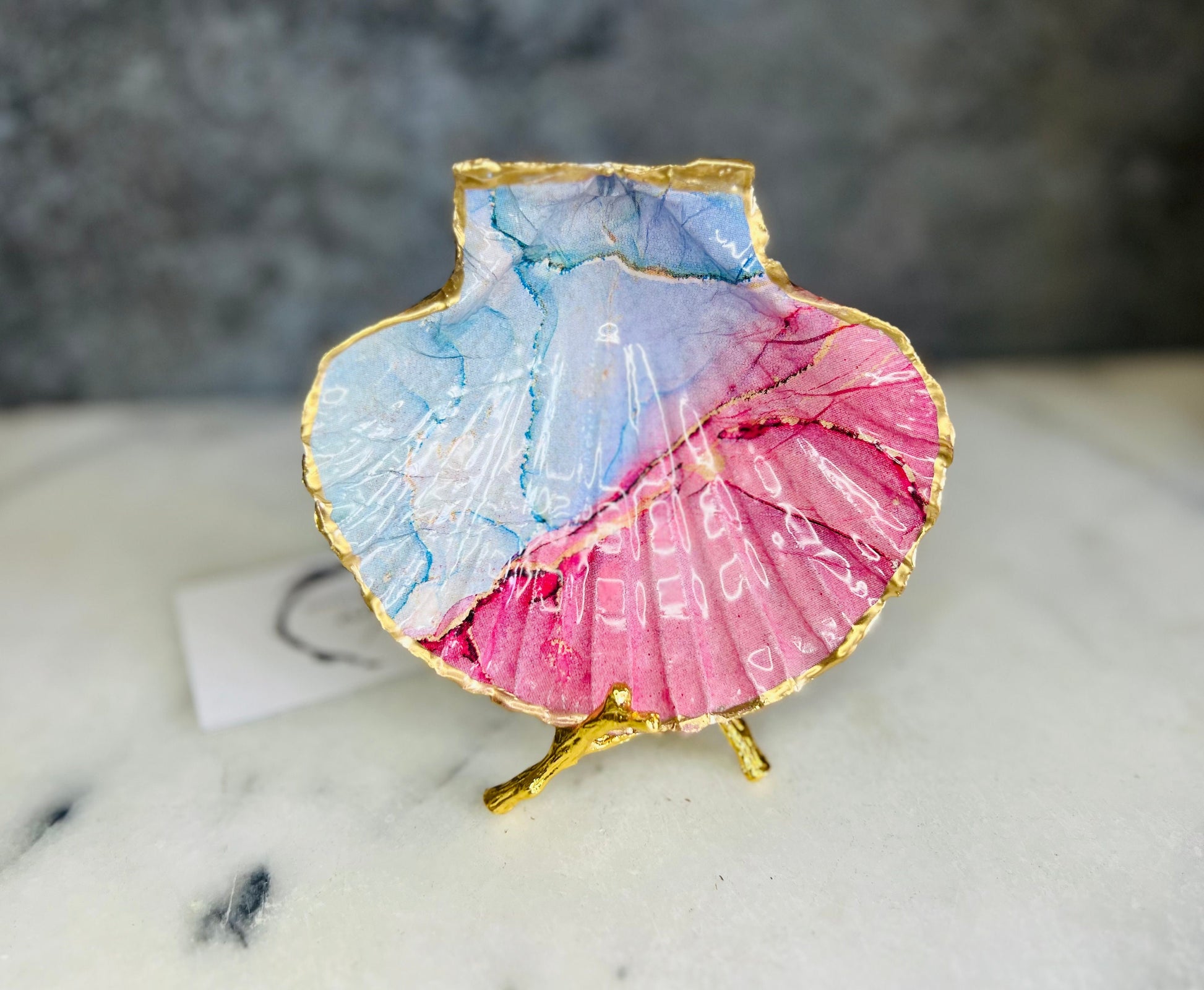 A handmade scallop shell trinket dish painted in soft pink and pale blue tones in a flowing, watercolor-style pattern, finished with a gold leaf edge and sealed with a glossy clear resin. The natural shell shape is visible, with the ridged texture of the scallop shell showing through the design. The dish is shown on a light surface and works as a small decorative tray suitable for holding rings or jewellery in a bedroom or bathroom.